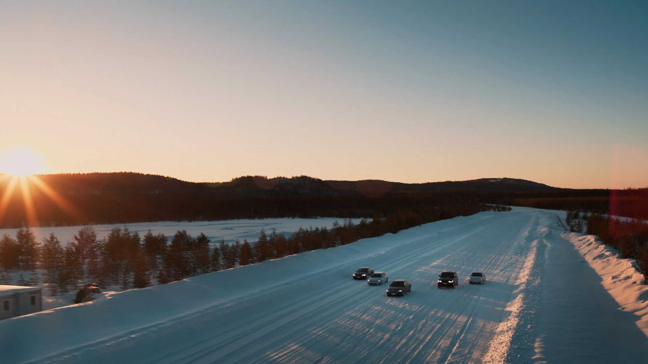 Snow-covered road with six parked cars, leading towards a tree-lined horizon with mountains in the distance, during sunset.