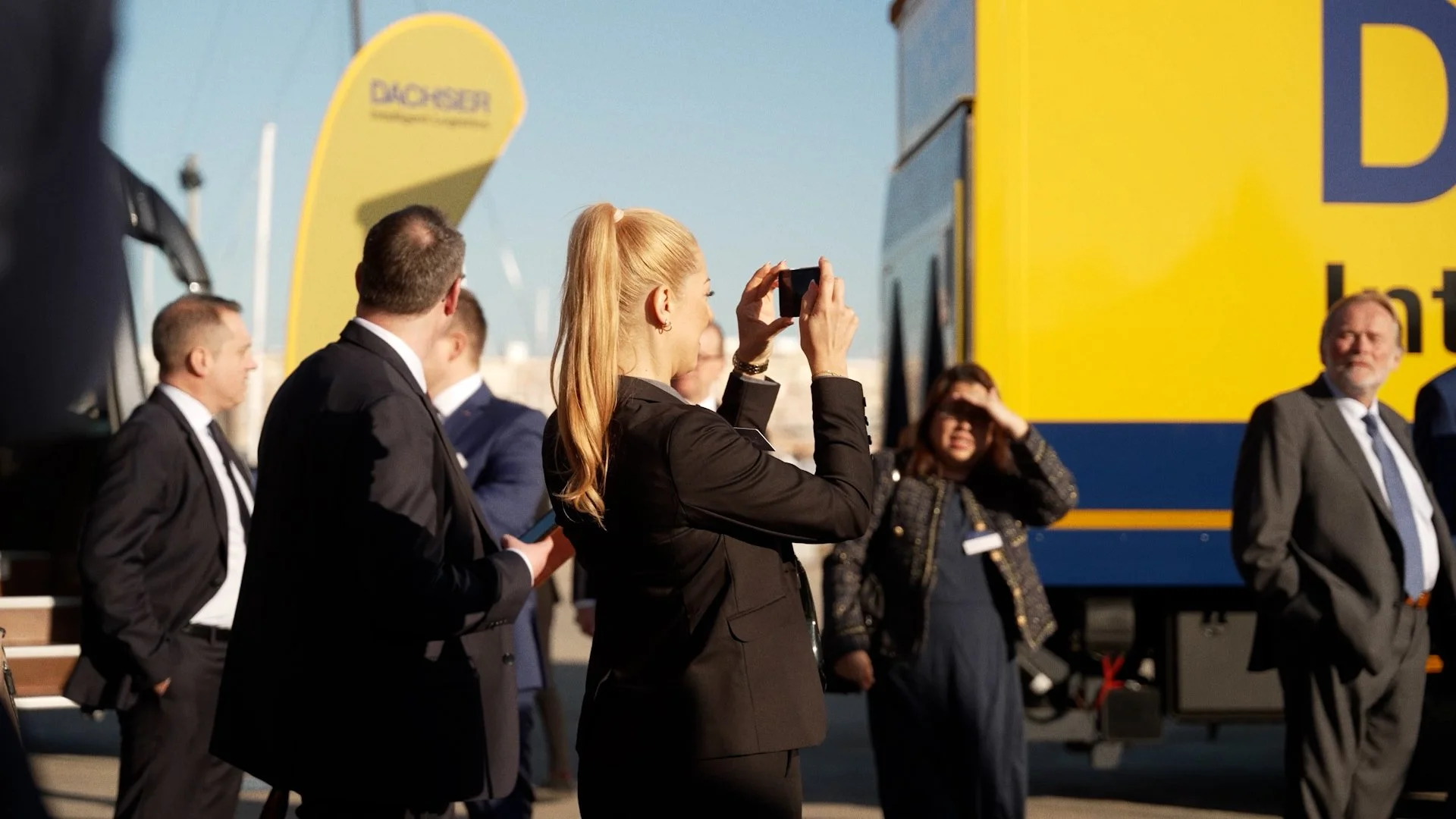 A woman with red hair taking a photo with a smartphone at an outdoor event, with several men in suits and a yellow and blue bus in the background.