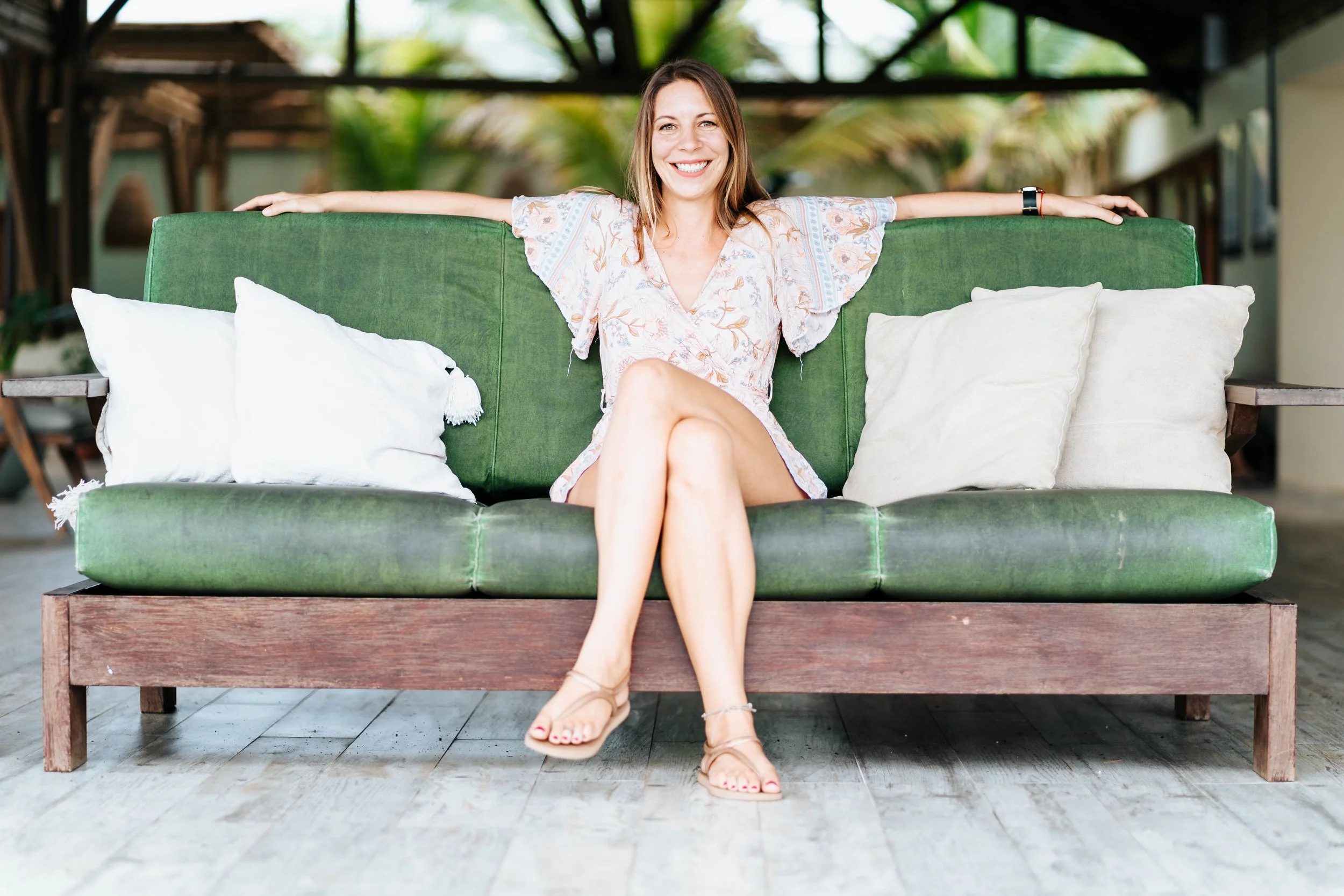 Smiling woman sitting on a green outdoor sofa with white pillows, arms stretched out along the backrest, cross-legged, wearing a floral dress and sandals, with a tropical background.