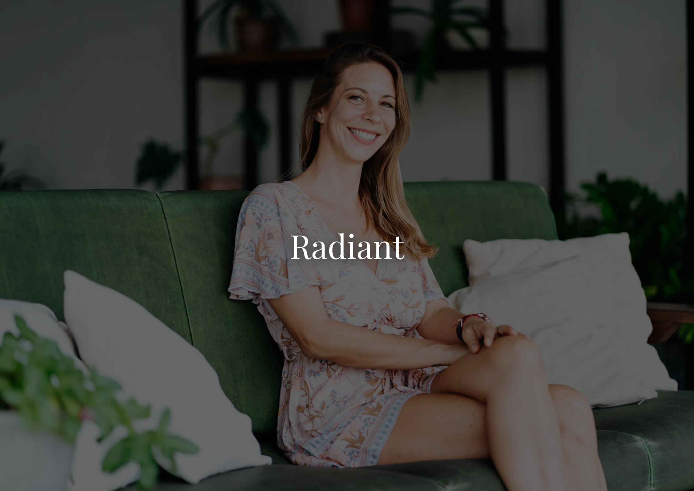 A woman with long brown hair smiling while sitting on a green couch, wearing a light-colored floral dress, with houseplants and a black shelving unit in the background.