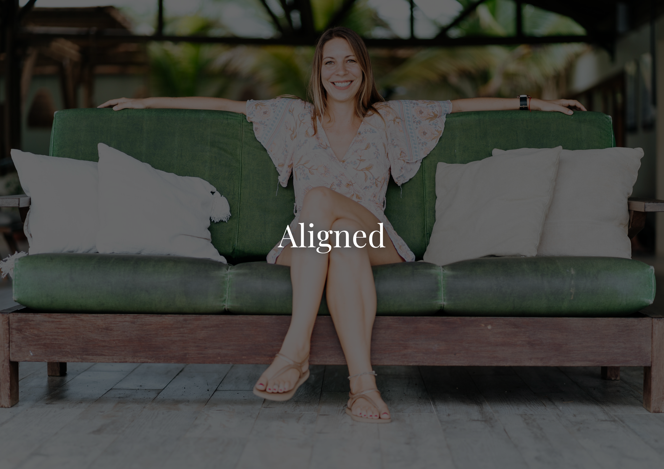 A woman with long brown hair smiling, sitting on a green outdoor sofa with three white pillows, wearing a light patterned dress and sandals, with her arms spread on the sofa backrest, arms extended, in a tropical or outdoor setting.