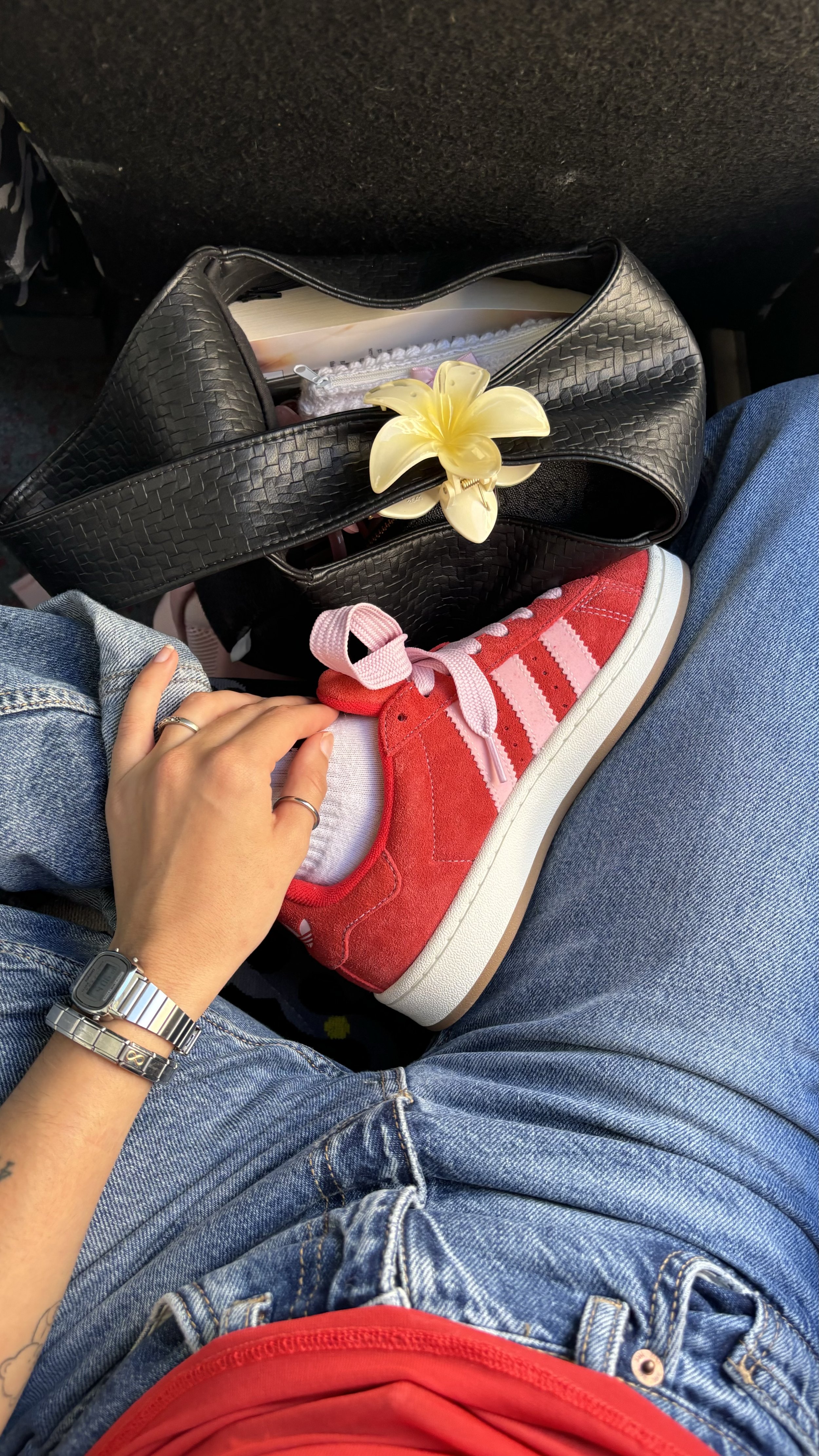Person sitting on an airplane with their legs crossed, wearing red Adidas sneakers with pink laces and blue jeans. The person's left hand, wearing a silver watch and rings, rests on their leg. A black purse with a yellow flower decoration and a book inside is beside them in the seat pocket.