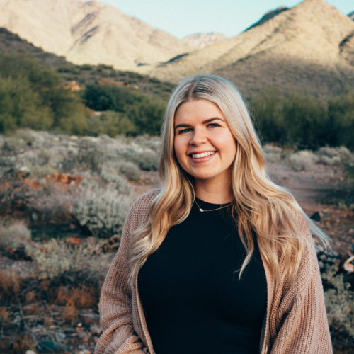 A smiling young woman with long blonde hair standing outdoors in a desert landscape with mountains in the background.