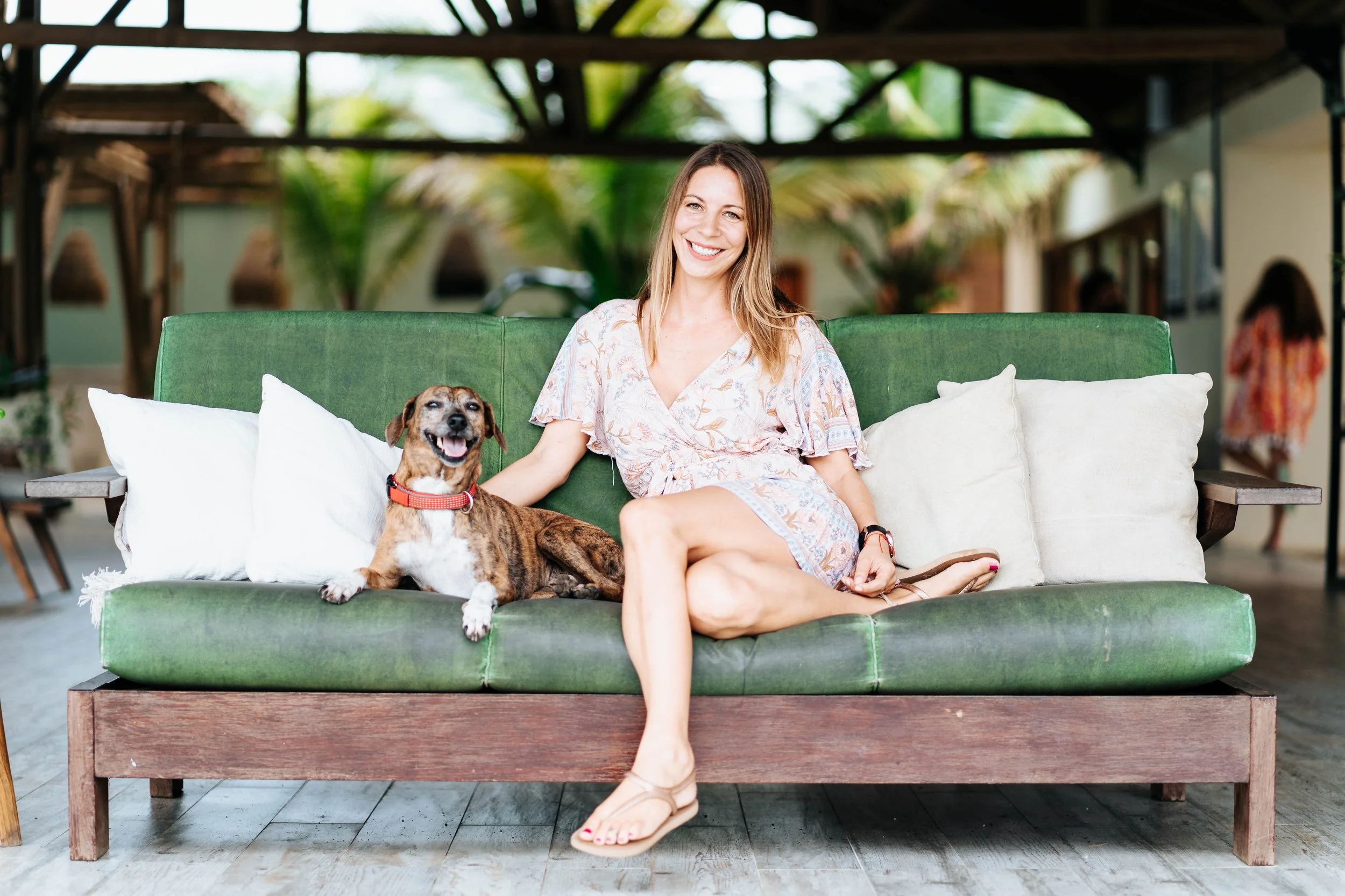 A woman sitting on a green outdoor sofa with a smiling dog beside her. The woman is wearing a light-colored dress and sandals, and the dog has a red collar. The background shows a covered outdoor area with palm trees and other people.