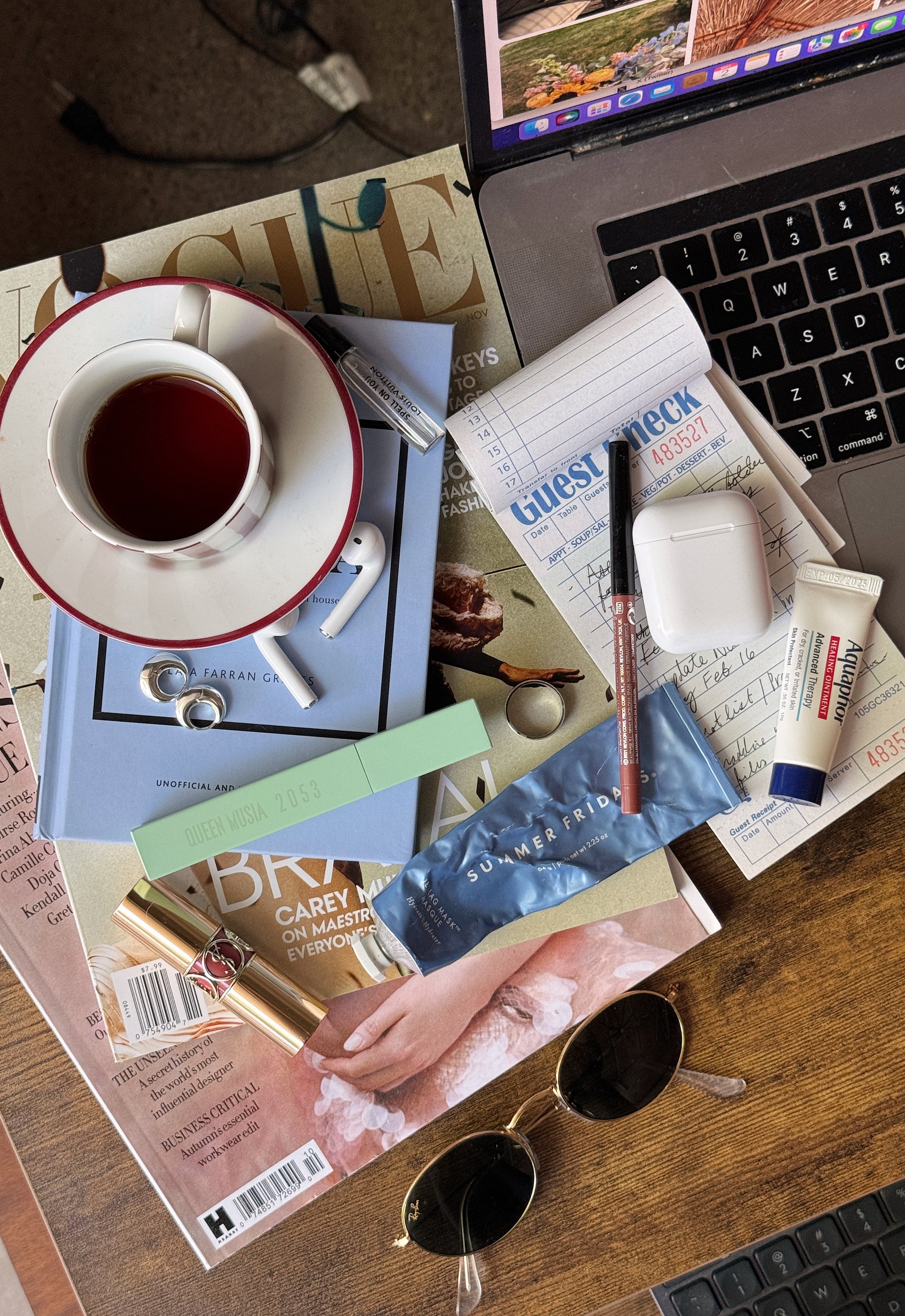 A cluttered desk with a laptop, a cup of tea on a saucer, a notepad, a pen, lipstick, a tube of hand cream, a pair of sunglasses, magazines, and various papers.