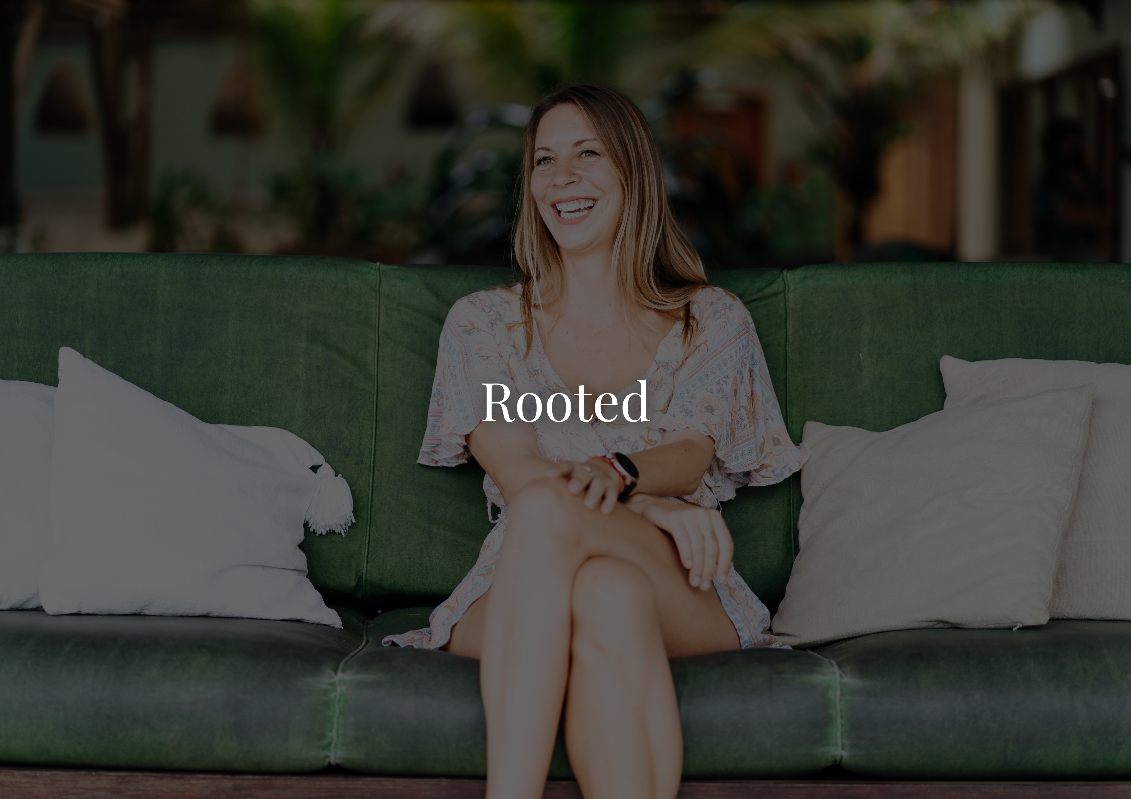 Smiling woman sitting on a green couch with white pillows, wearing a patterned dress and a smartwatch, in a cozy indoor setting.