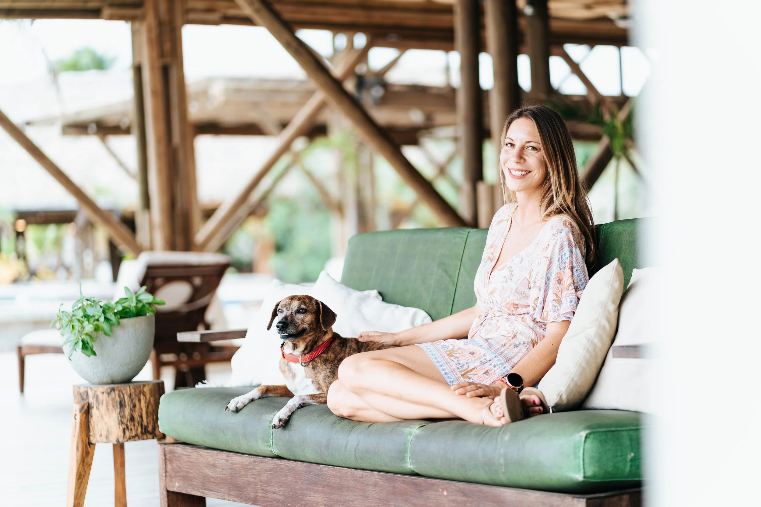 A smiling woman sitting on a green outdoor sofa with a dog, in a rustic wooden structure with greenery outside.