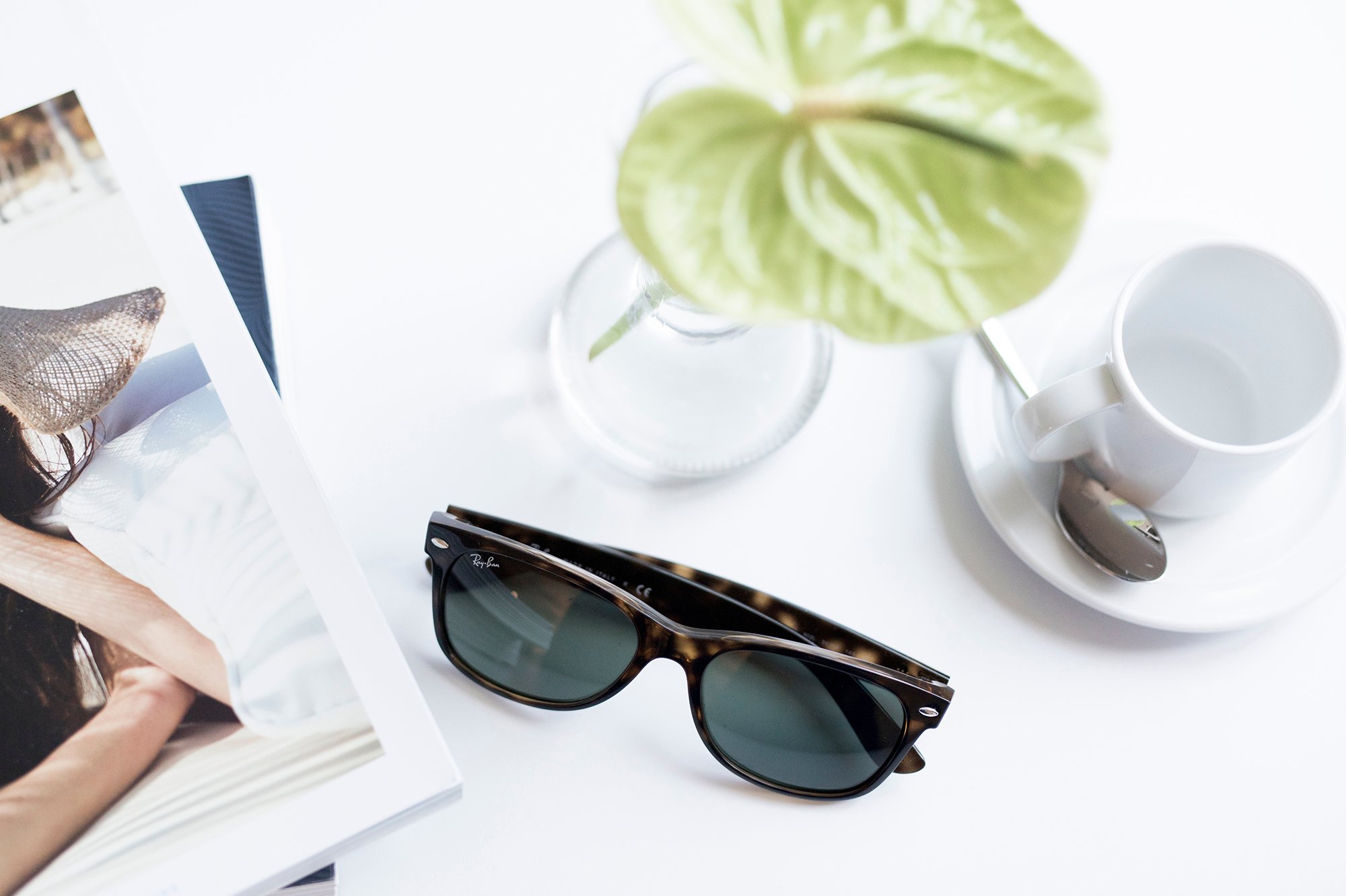 Turtle shell sunglasses, a magazine, a potted green plant, a white coffee cup with a saucer and spoon, and a small jar on a white surface.