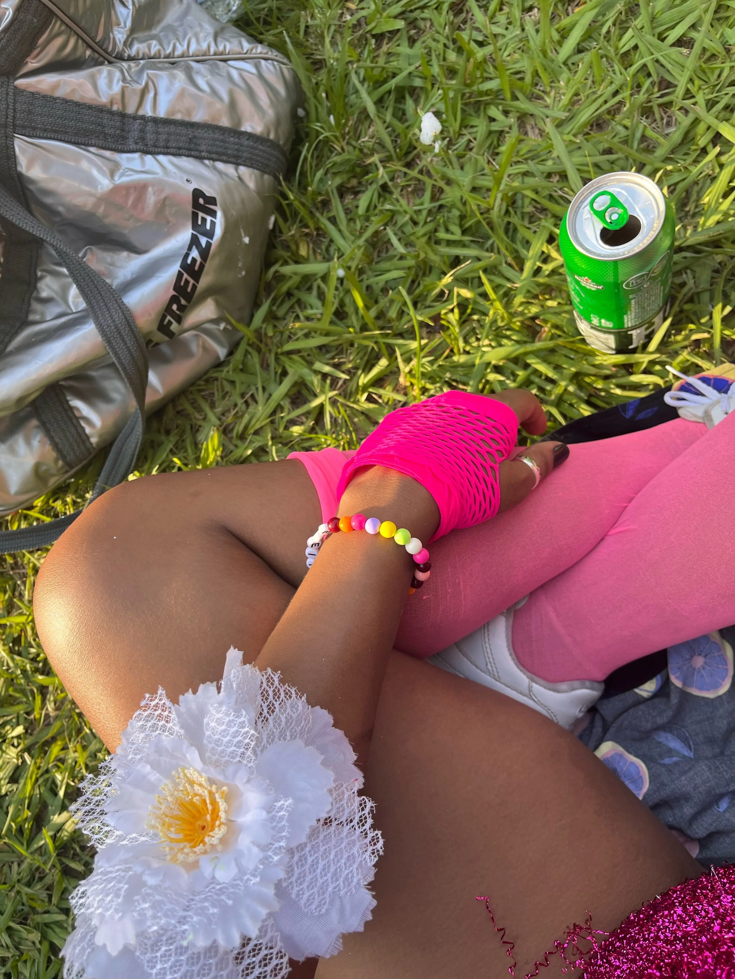 Close-up of a person sitting on grass, wearing pink clothing, a white flower accessory on the thigh, and a colorful beaded bracelet, with a soda can and a backpack nearby.