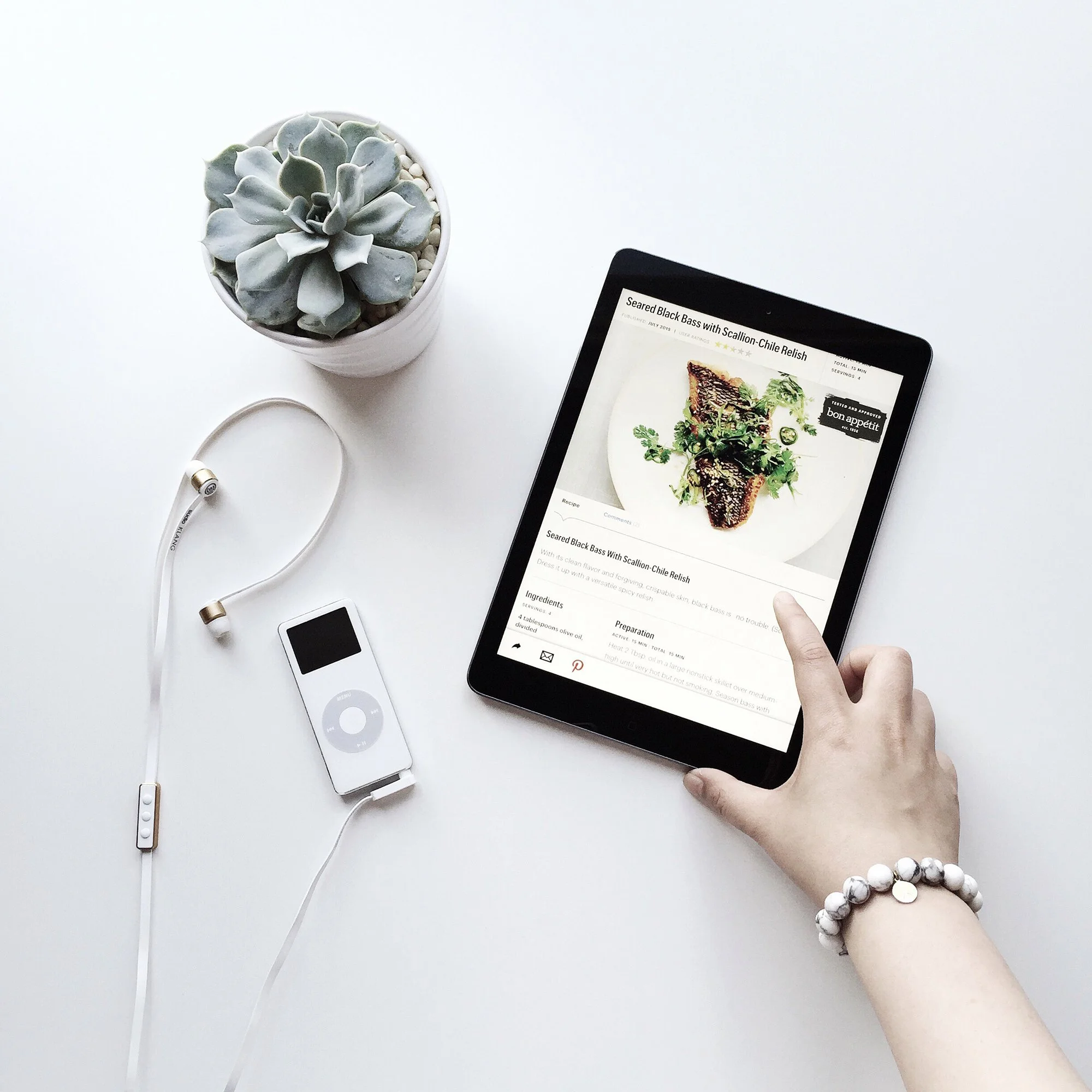 Person using a tablet to view a recipe for seared black bass with scallion-chile relish, with a potted succulent plant, white earbuds, and an iPod on a white surface.