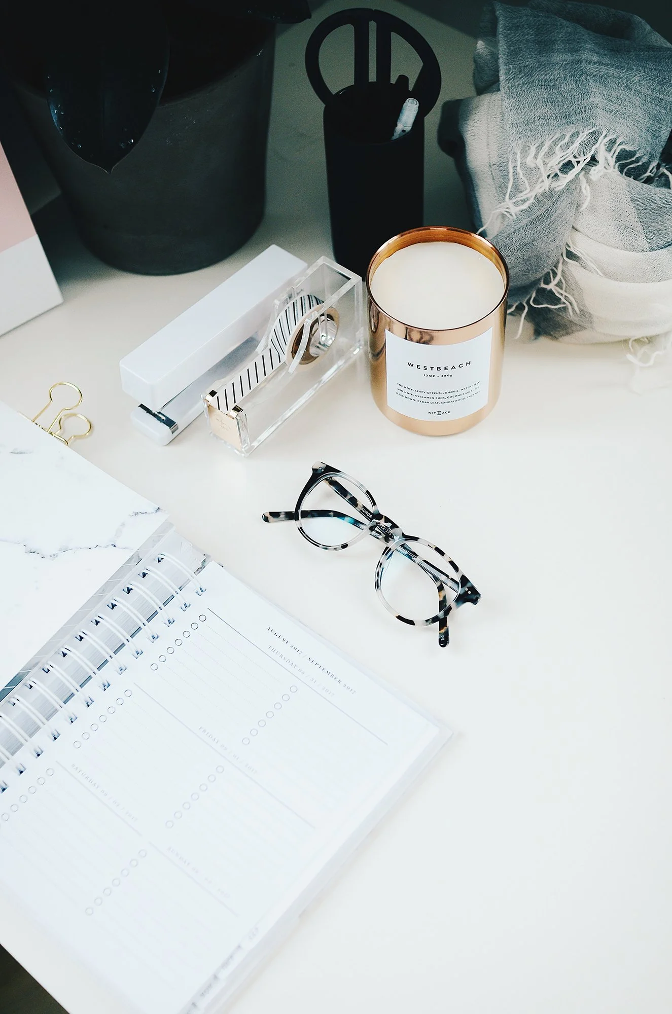 A desk with a pair of glasses, a planner, a candle, a container with pens, and some office supplies.