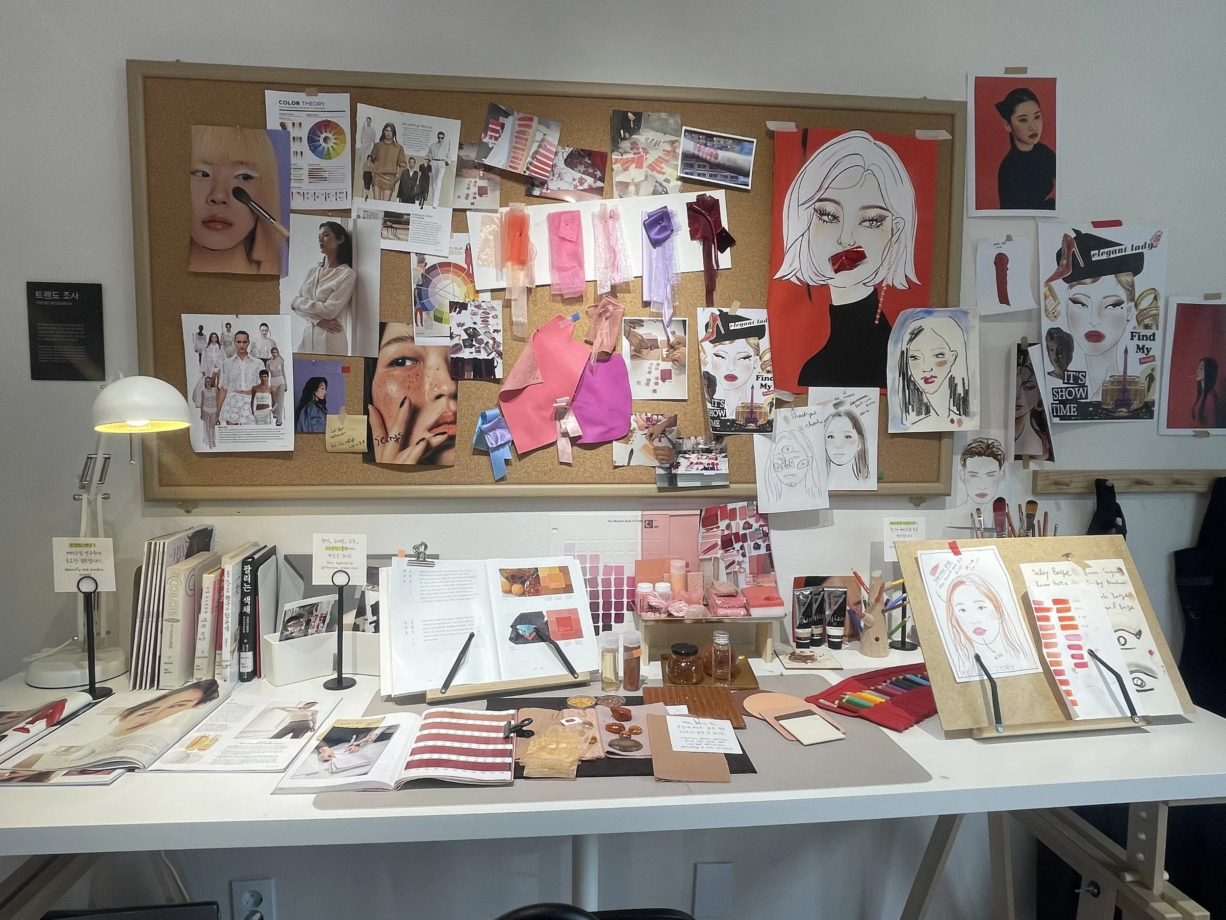Creative workspace with mood boards, fashion sketches, fabric swatches, color palettes, and art supplies on a white table and a corkboard filled with fashion and artistic references.