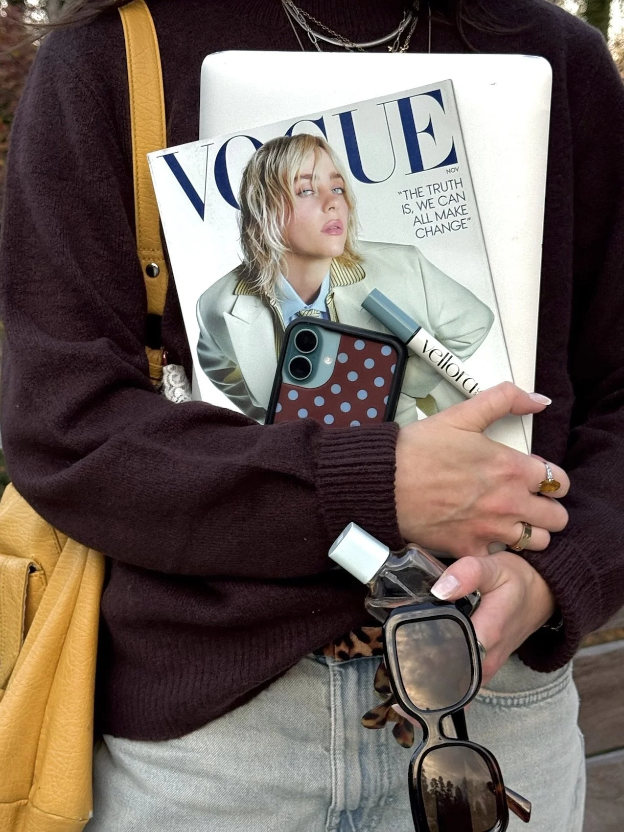 A person holding a Vogue magazine featuring a blonde woman on the cover, along with a phone, sunglasses, a hand sanitizer, a lip balm, and a yellow purse, while wearing a dark sweater and multiple necklaces.