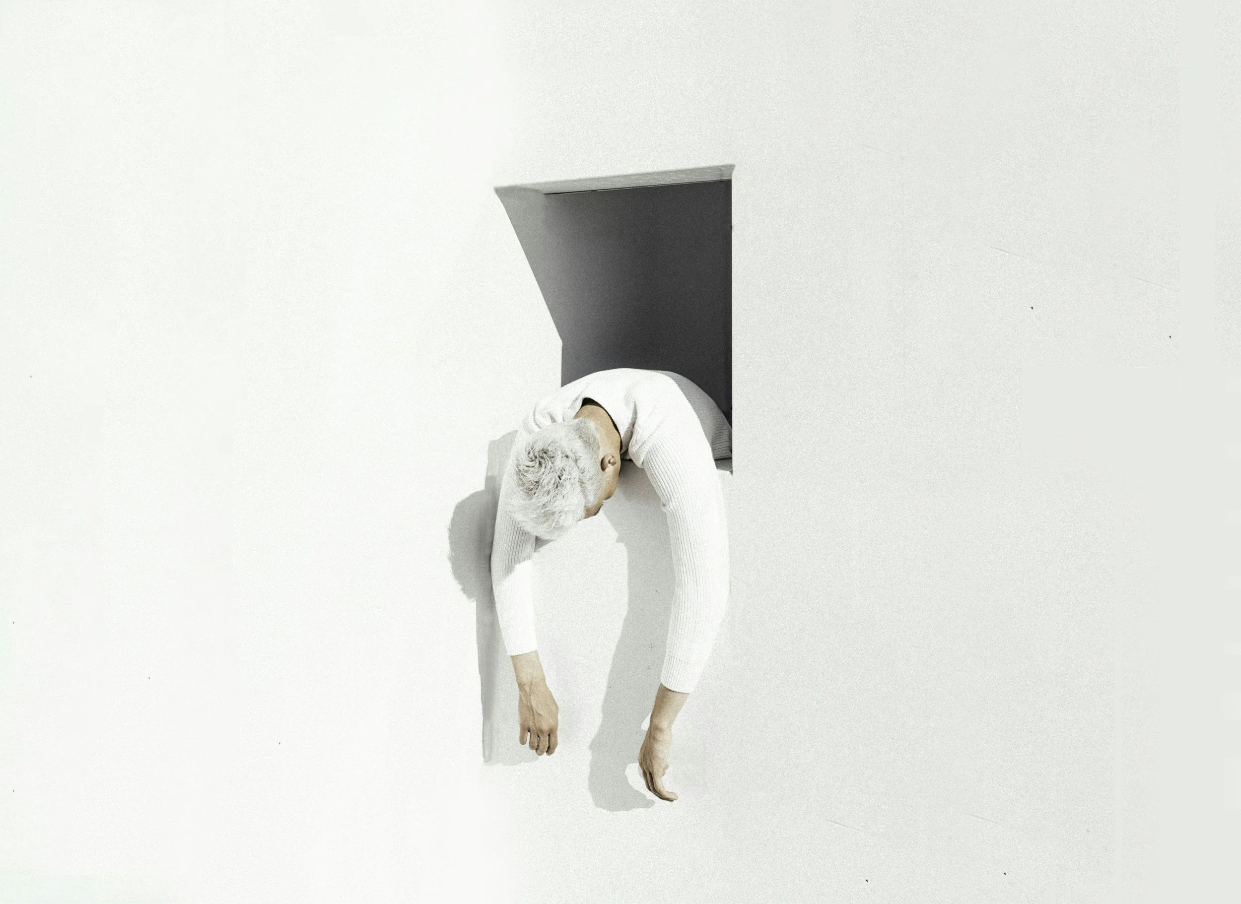 Person with white hair and a white sweater hanging upside down from a small square opening in a white wall