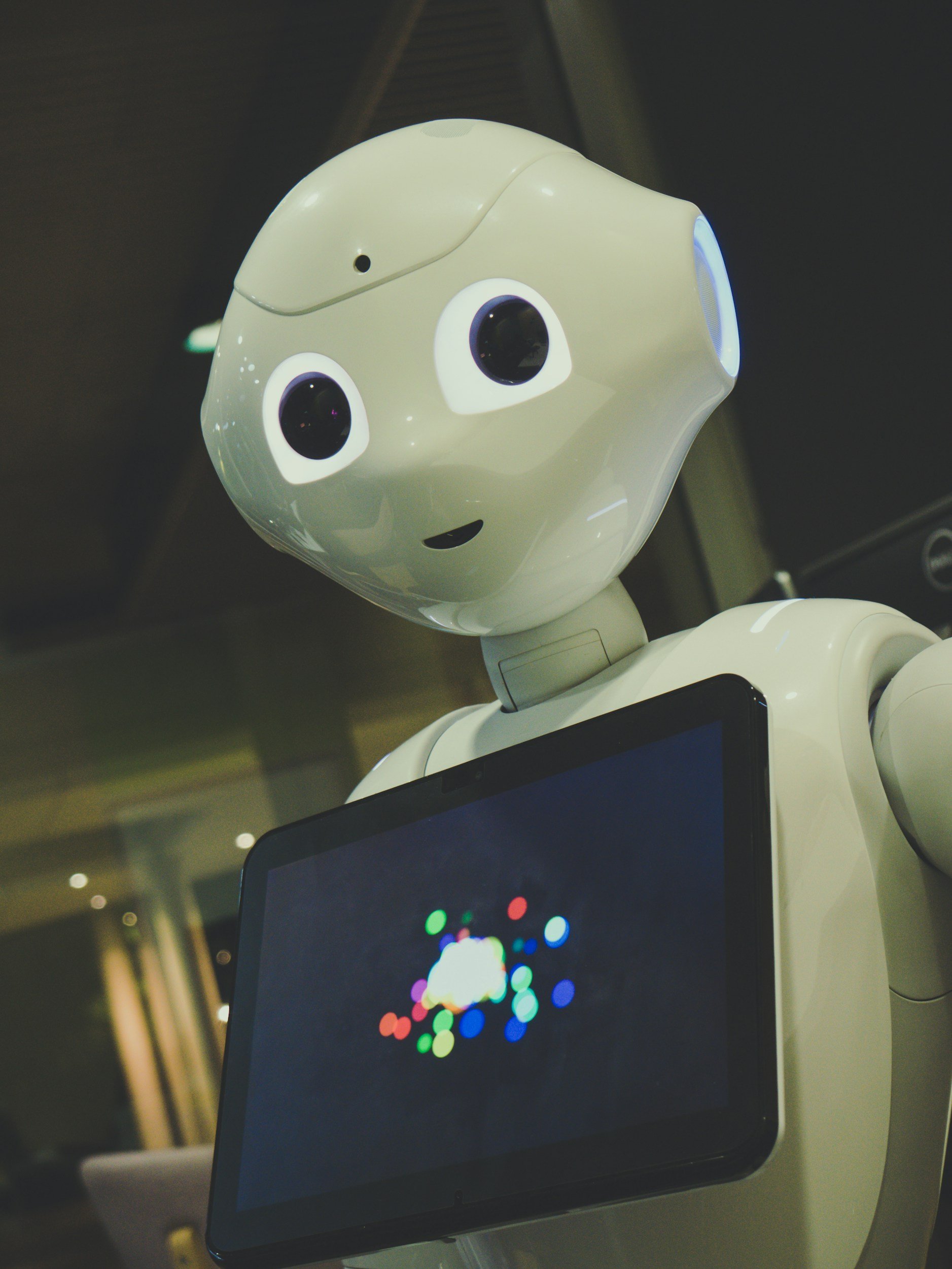 A robot with a large, rounded head and big expressive eyes stands in front of a laptop with a colorful spinning logo on the screen.
