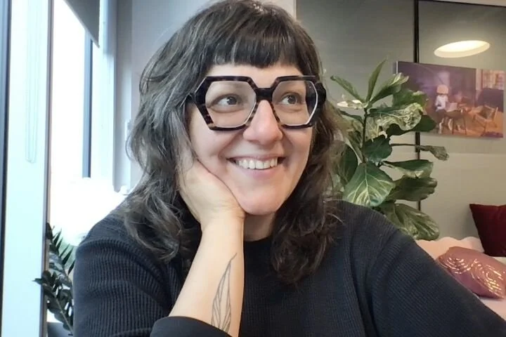 A woman with shoulder-length brown hair, wearing large black glasses and a black top, smiling and resting her face on her hand in an indoor setting with plants and artwork in the background.
