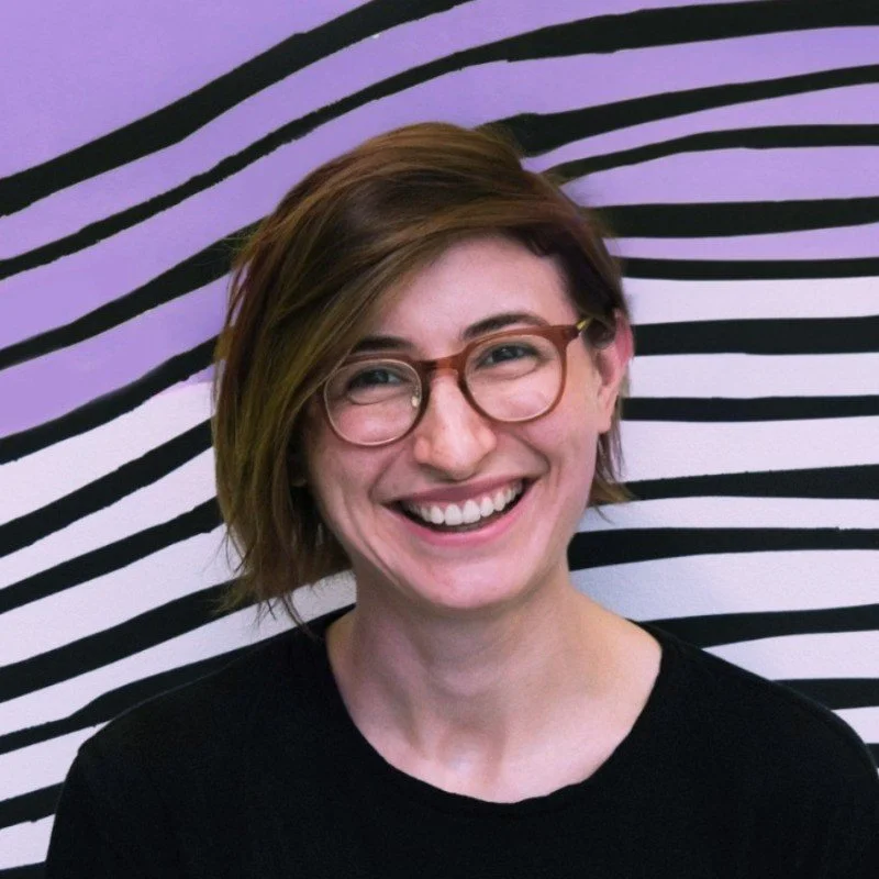 A woman with short brown hair, glasses, and a black shirt smiling in front of a black and white striped mural with curved lines.