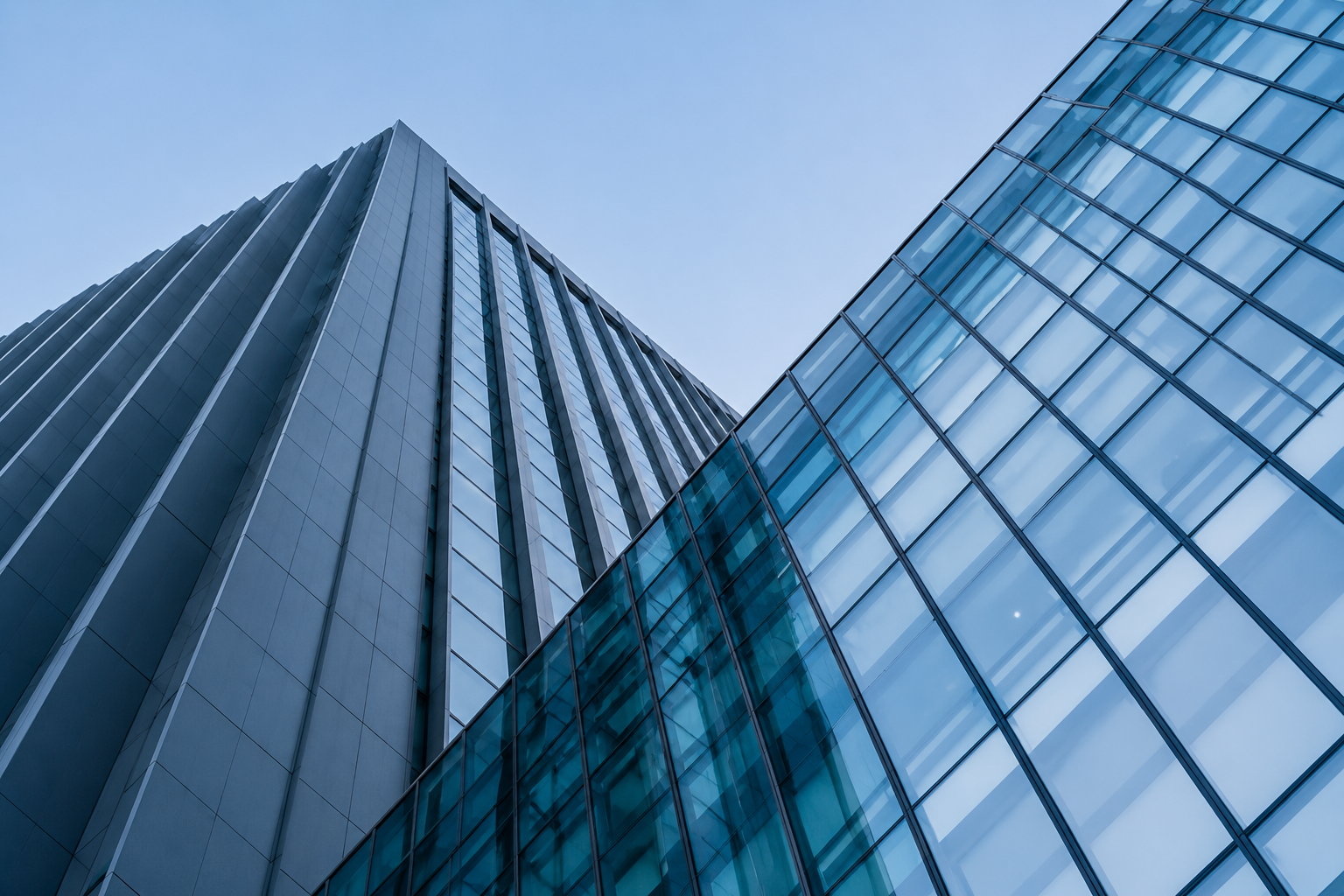 Two modern office skyscrapers with glass facades seen from below against a light blue sky.