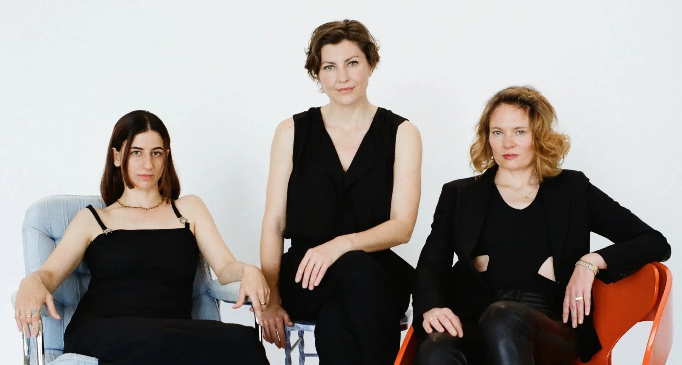 Three women sit together against a plain white background, wearing black outfits and looking directly at the camera.