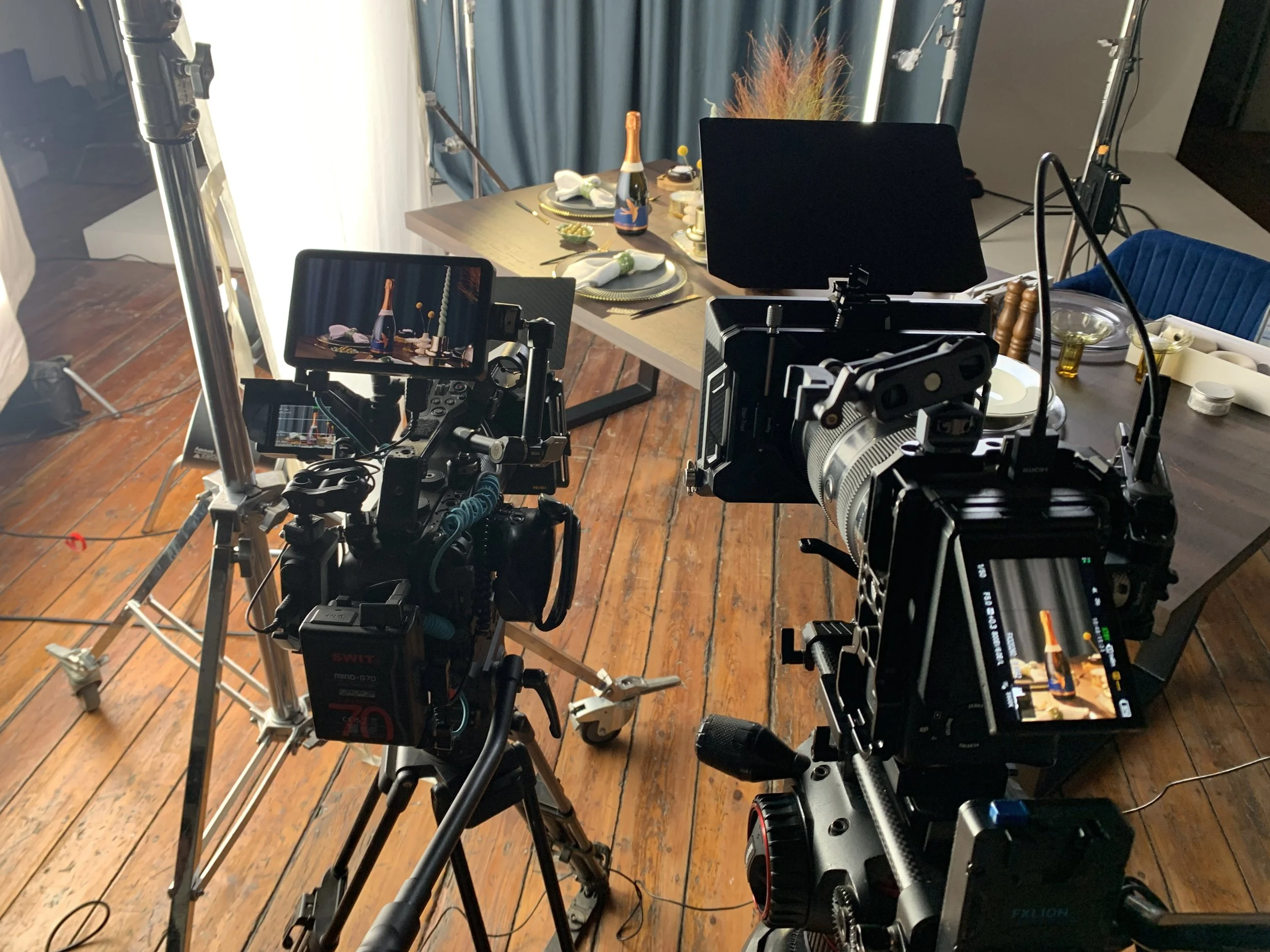 Two professional cameras on tripods filming a table set with dinnerware, champagne, and decorative items in a studio setting.