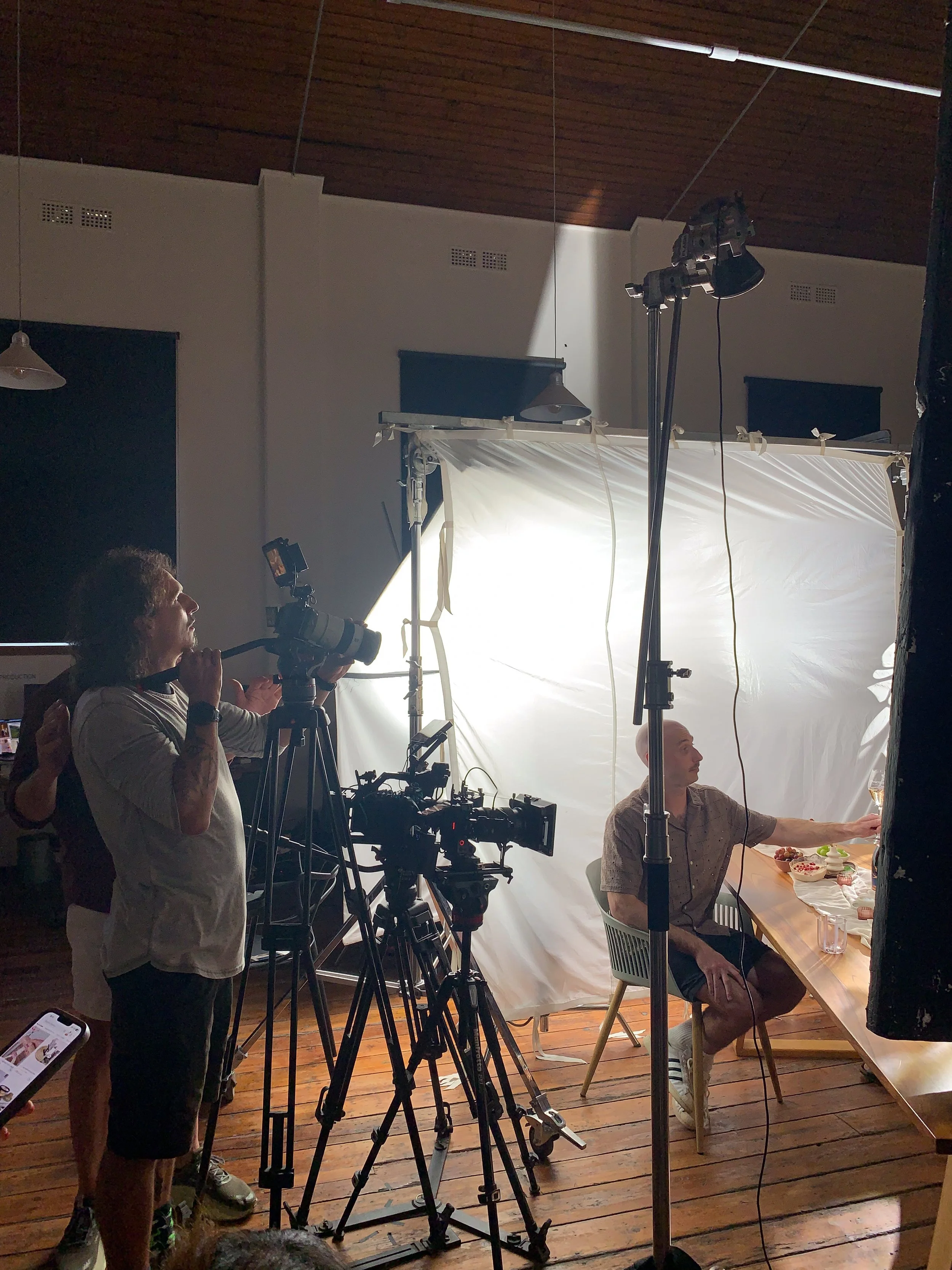A man sitting at a table with food, being filmed by a camera crew with professional lighting and equipment in a room with wooden floors and a white backdrop.