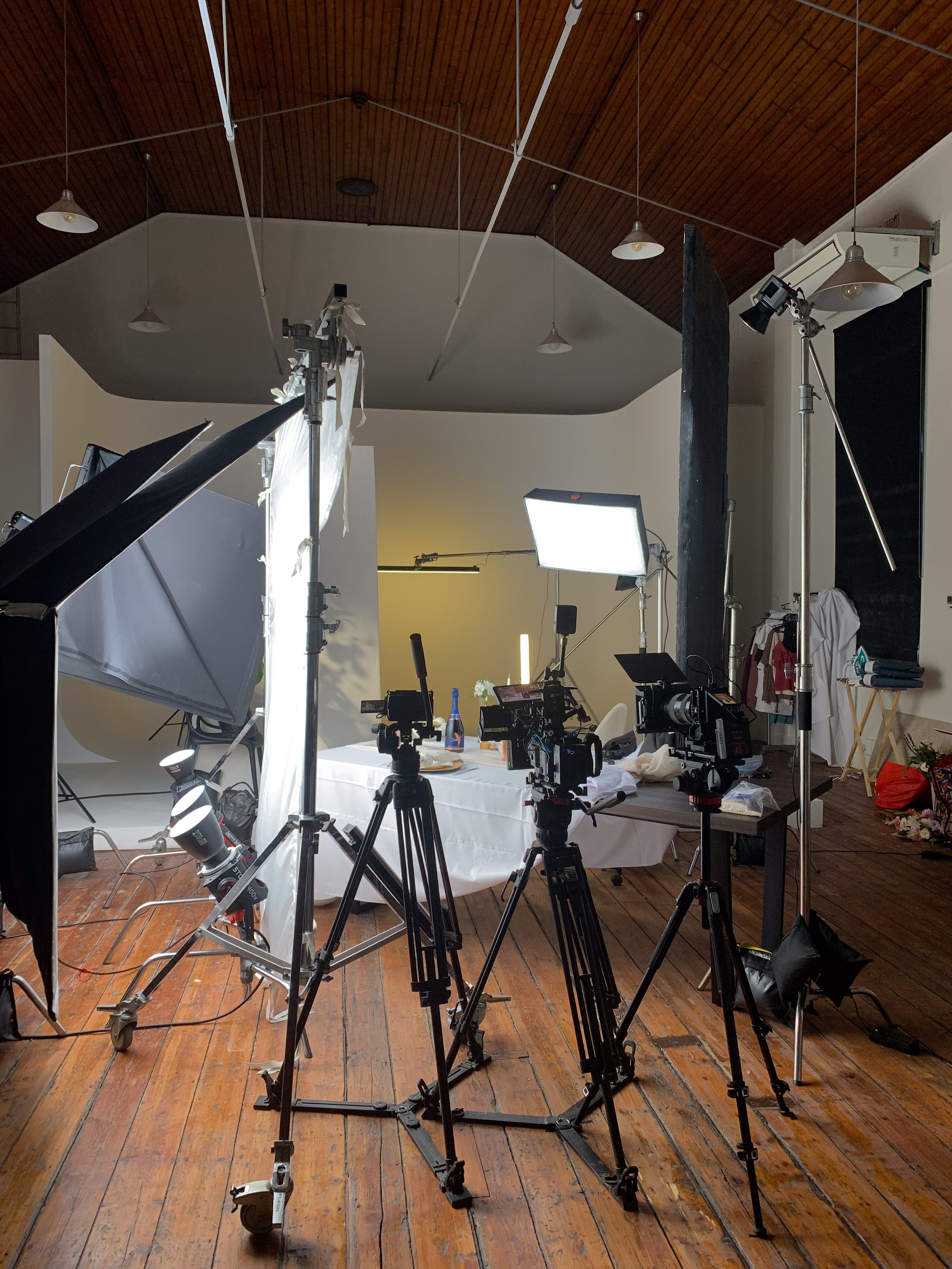 Photography studio setup with cameras, lights, and a table with items, including a bottle and flowers, in a room with wooden floor and ceiling.