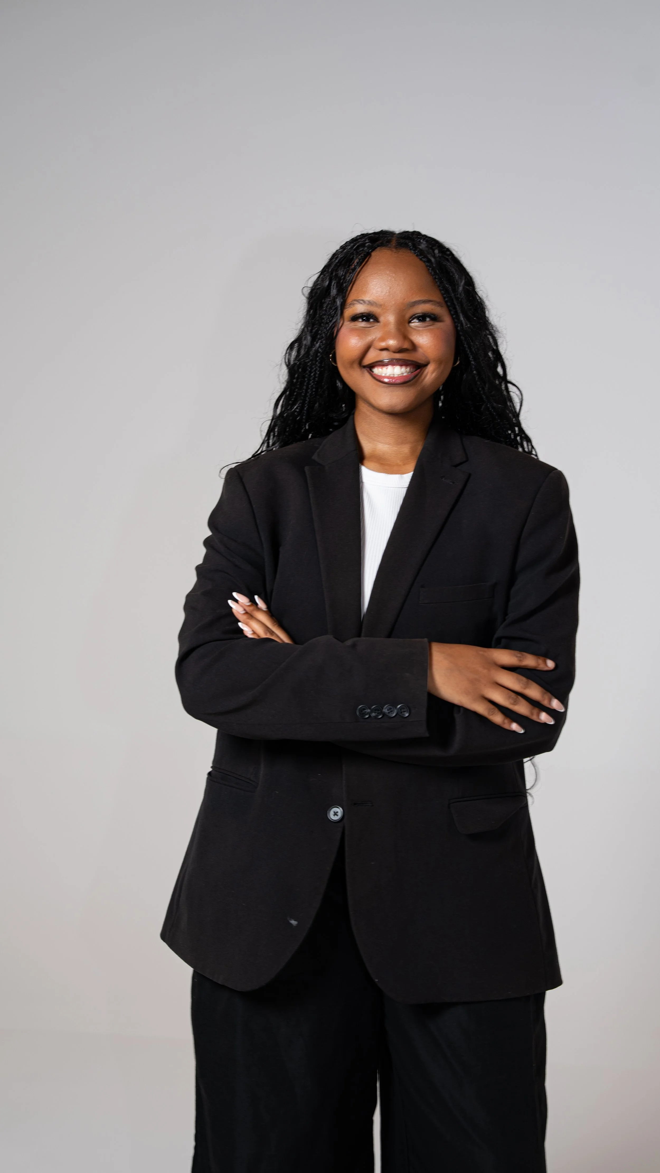 A confident young woman in a black suit with arms crossed, smiling against a plain gray background.