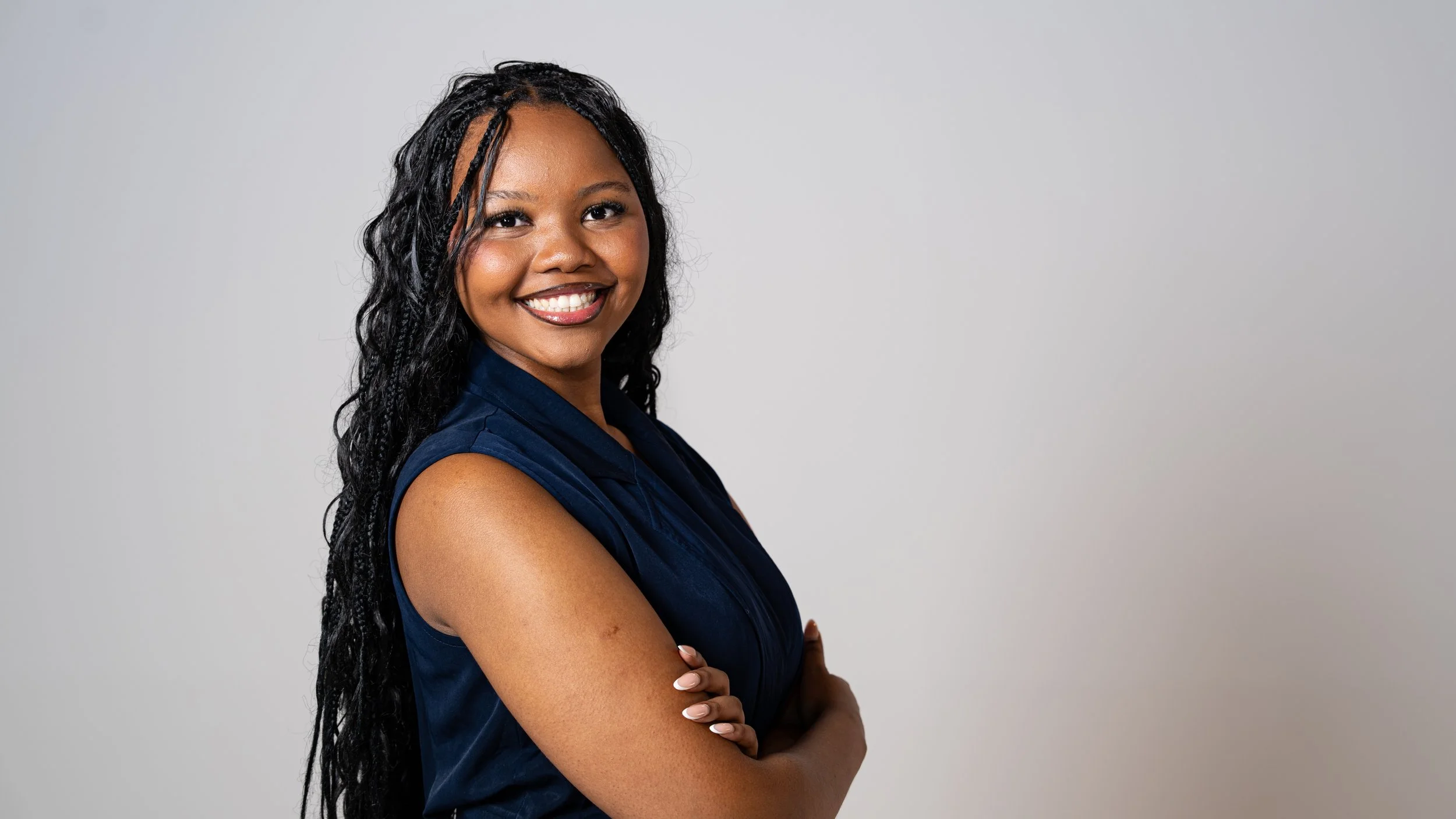 Portrait of a smiling woman with long curly black hair, wearing a sleeveless navy blue top, standing with her arms crossed against a light gray background.