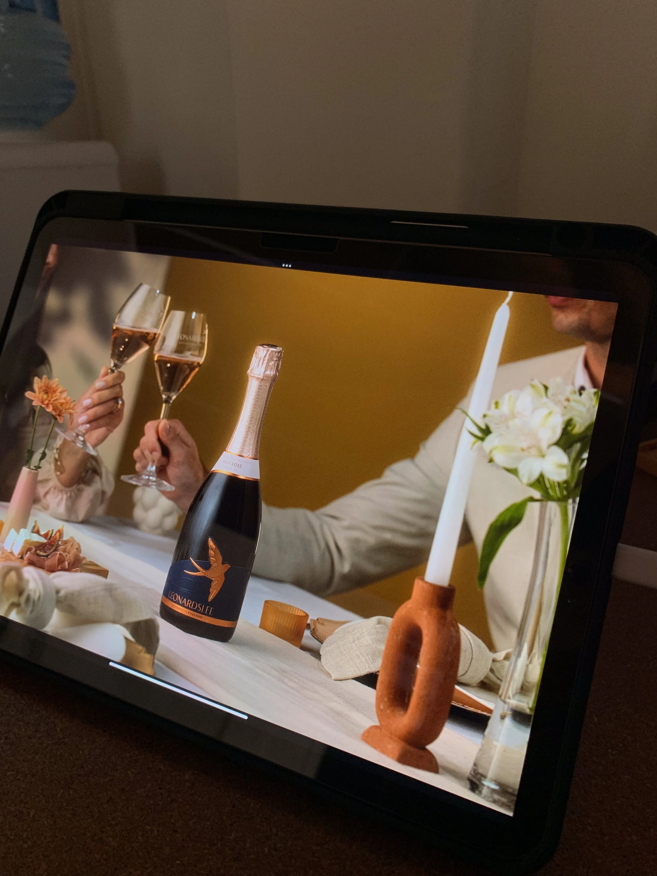 Tablet displaying a social gathering with people holding glasses of rosé wine, a bottle with a bird illustration, a candle, flowers, and table decorations.