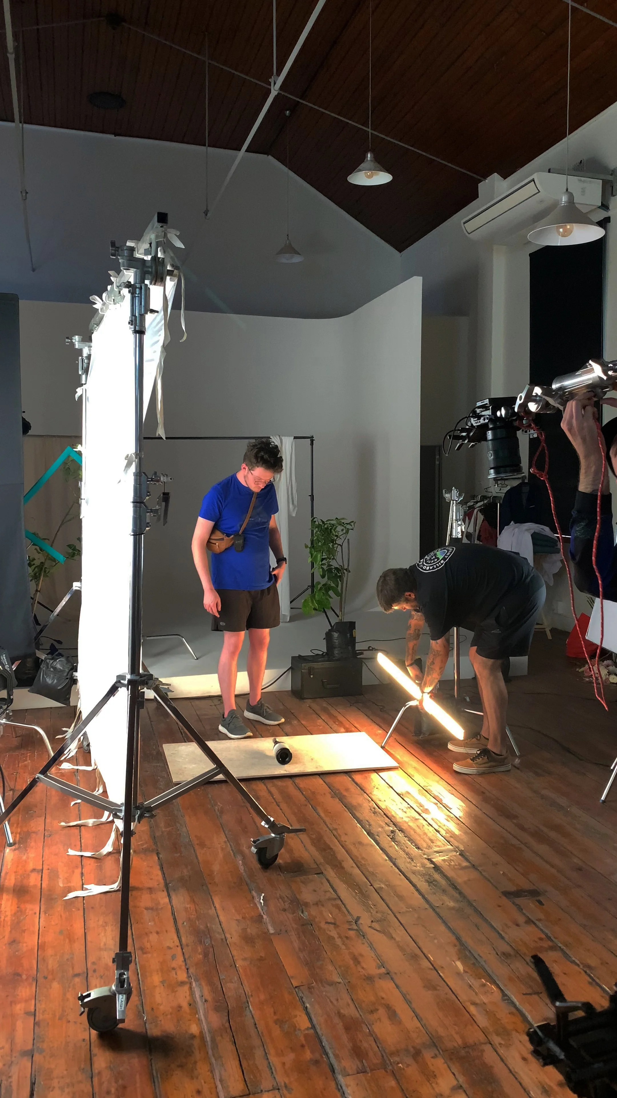 Filmmakers working on a scene with lighting equipment, a white backdrop, and green plants, indoors with wooden floors and ceiling.