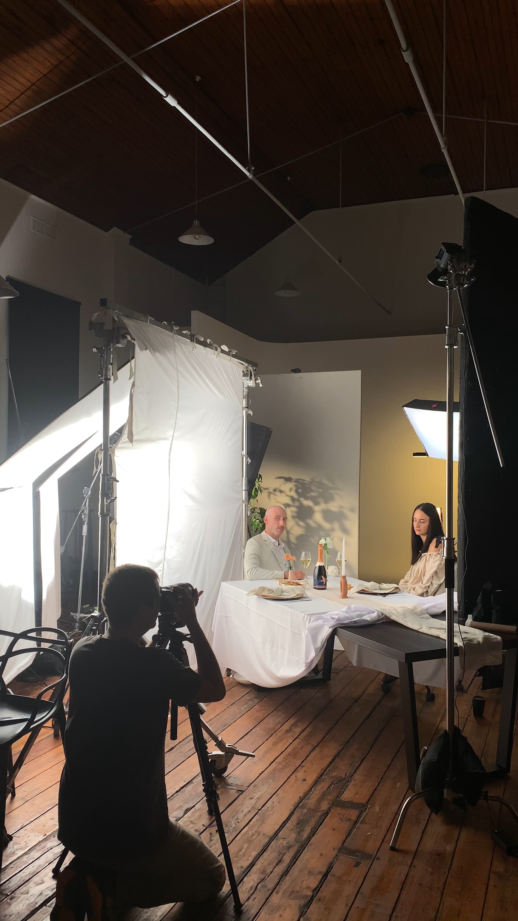 Filming of a faux wedding scene with two actors seated at a table, a photographer, lighting equipment, and a backdrop in an indoor studio with wooden floors and ceiling.