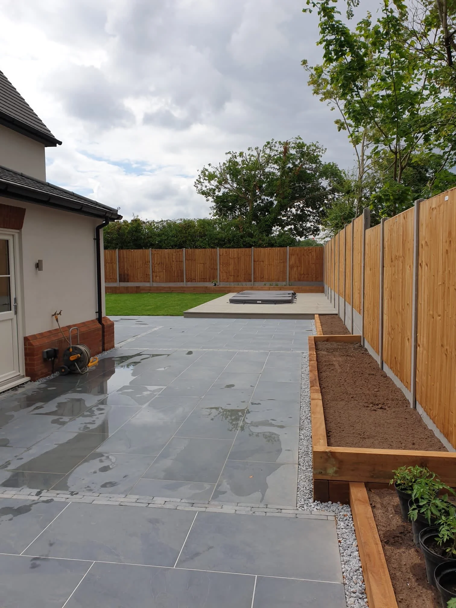 A backyard patio with wet grey tiles, a wooden fence, and newly planted flower beds along the right side. There is a house on the left with a door and brick accents. The sky is cloudy, and some trees are visible in the background.