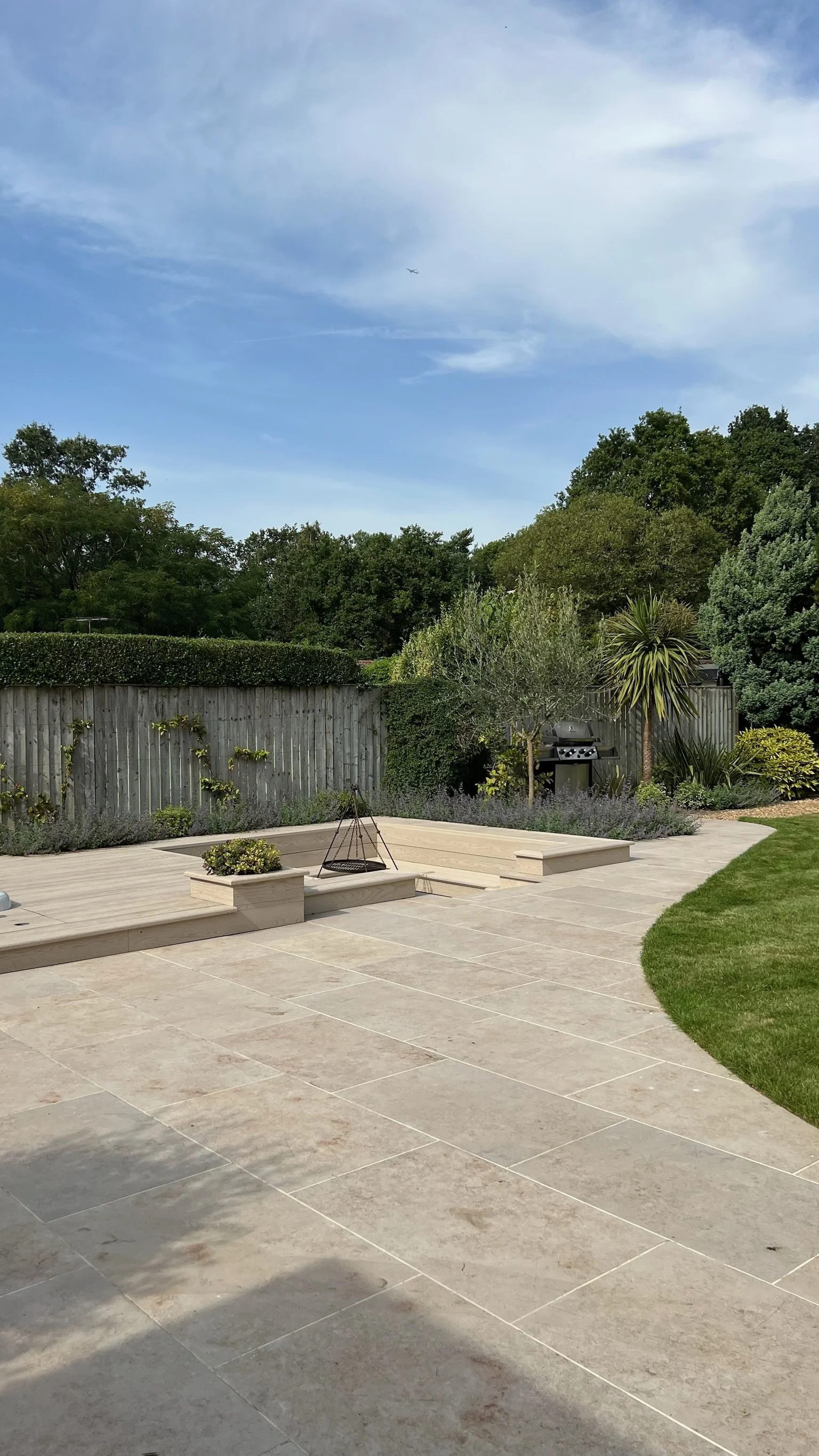 Garden with tiled patio, raised deck with fire pit, fencing, trees, bushes, and grill under a partly cloudy blue sky. Expert landscapers and garden designers in Surrey, Hampshire, Berkshire, Camberley, London and surrounding areas.