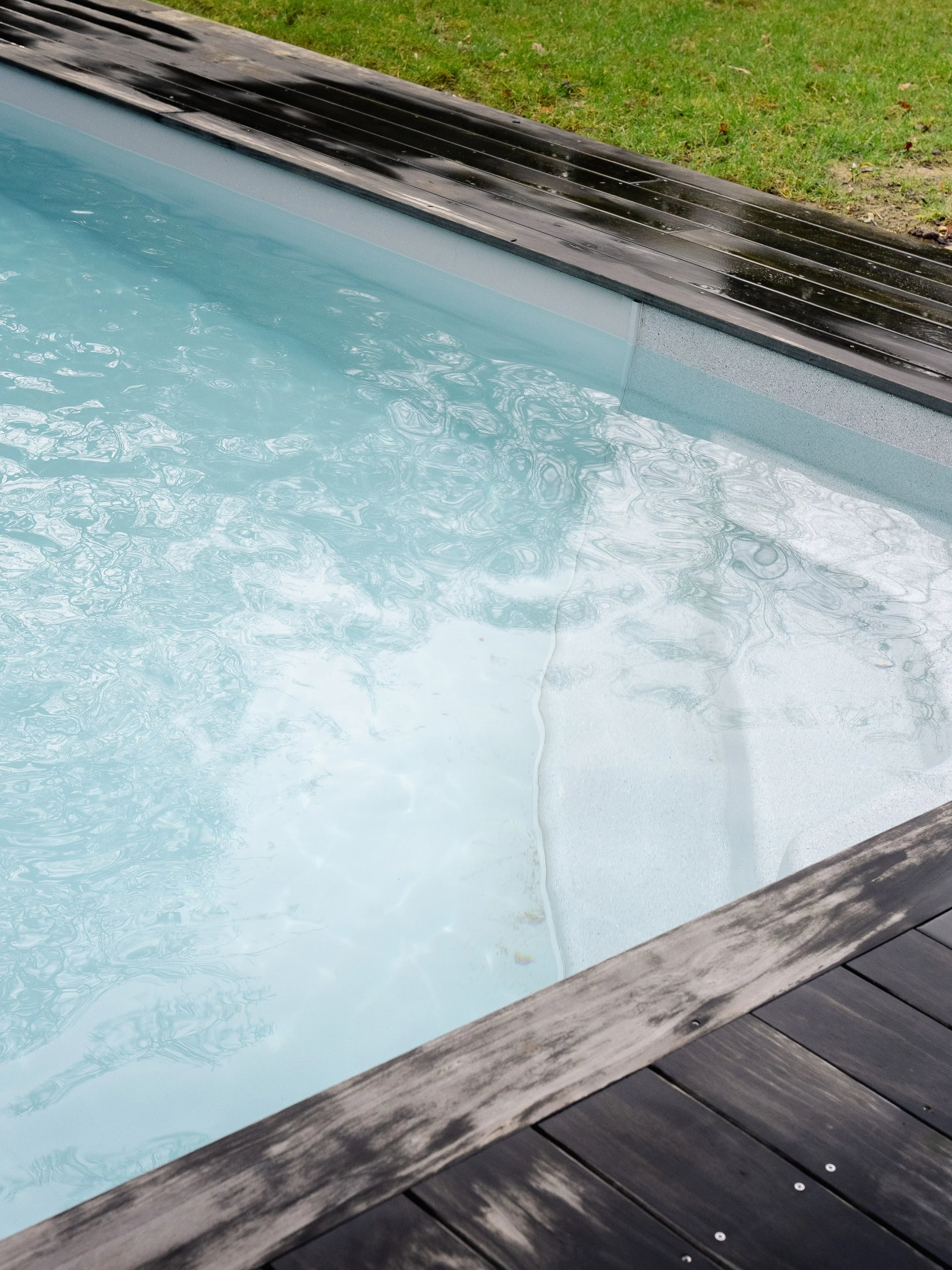 Close-up of a swimming pool with clean, clear water and a wooden deck border, with grass and some leaves in the background.