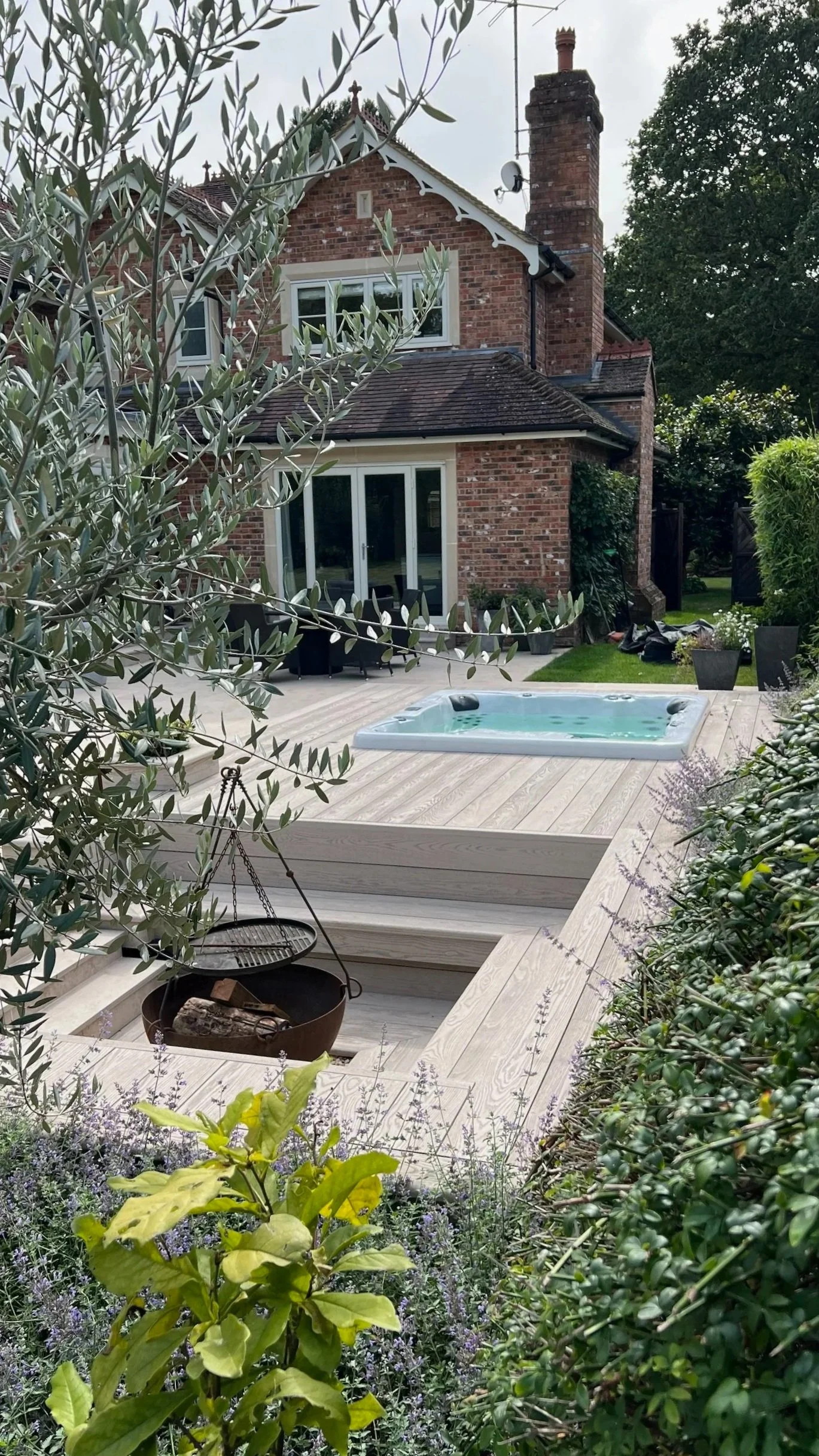 Garden with a hot tub on a raised wooden deck, firepit area, surrounded by lush green plants and bushes. In the background, there's a patio. Expert landscapers and garden designers in Surrey, Hampshire, Berkshire, Camberley, London.