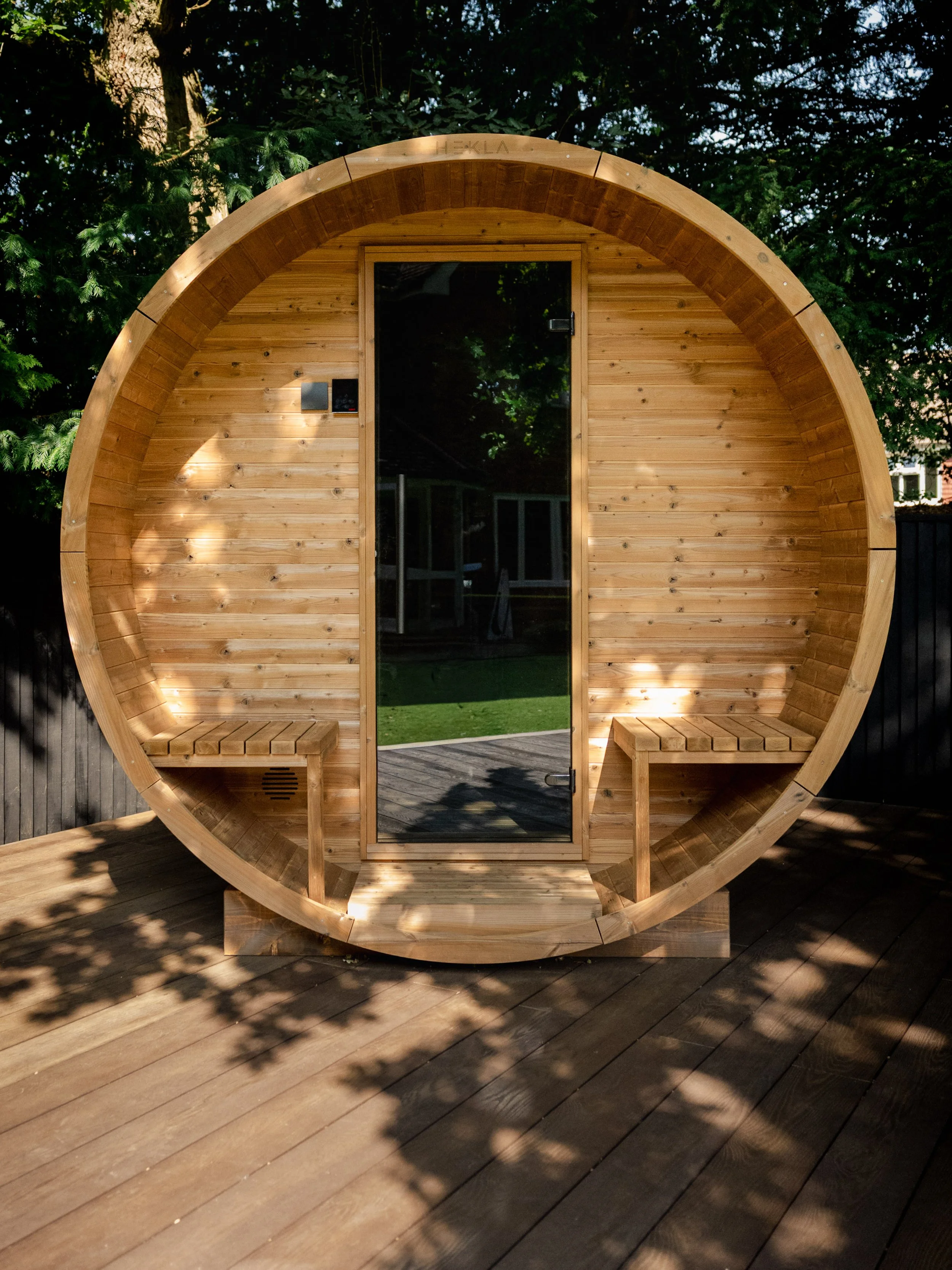 A round wooden sauna with a glass door, located outdoors on a wooden deck, surrounded by greenery and trees. Expert landscapers and garden designers in Surrey, Hampshire, Berkshire, Camberley, London and surrounding areas.