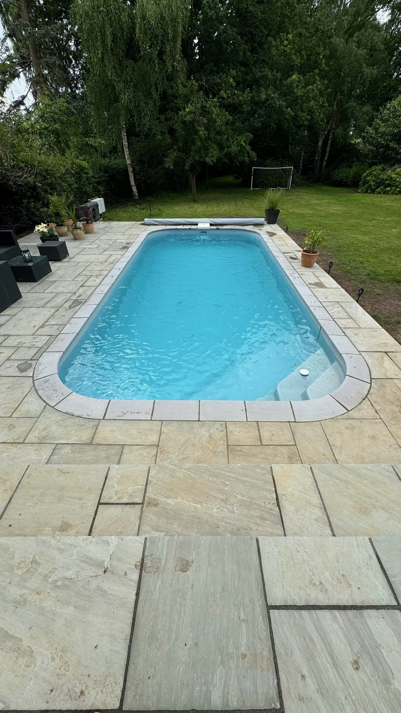 Backyard with a rectangular swimming pool, patio furniture, potted plants, and a grassy lawn with trees and a small soccer goal in the background.