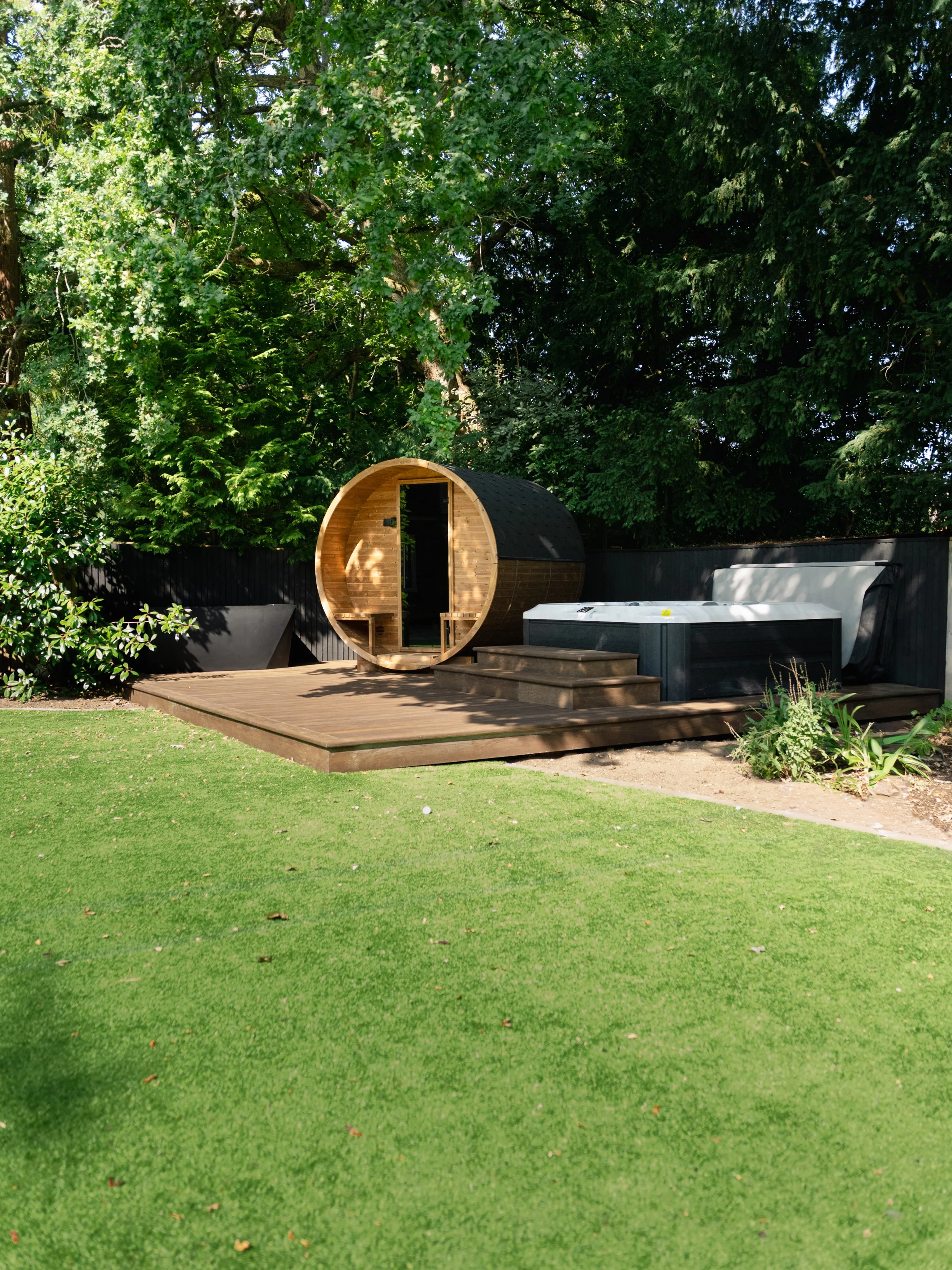 A backyard with a wooden deck and a hot tub, next to a small wooden sauna with a rounded door, surrounded by green trees and a grassy lawn.