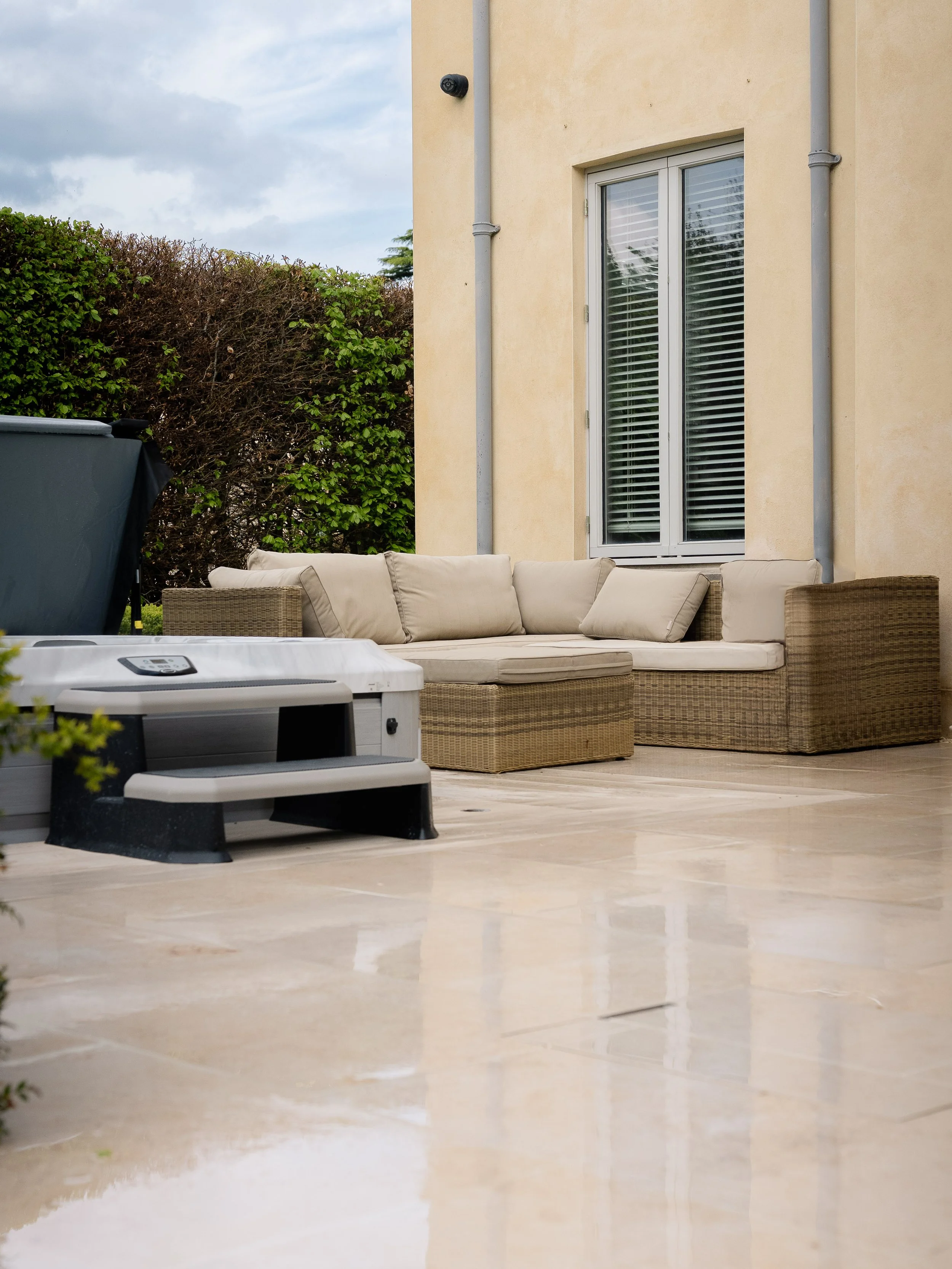 Outdoor patio with a beige sectional sofa, a small beige ottoman, a gray and black step stool, and a gray safety cover on a grill, next to a light yellow house wall with a window and a black security camera, with a hedge and cloudy sky in the backgro