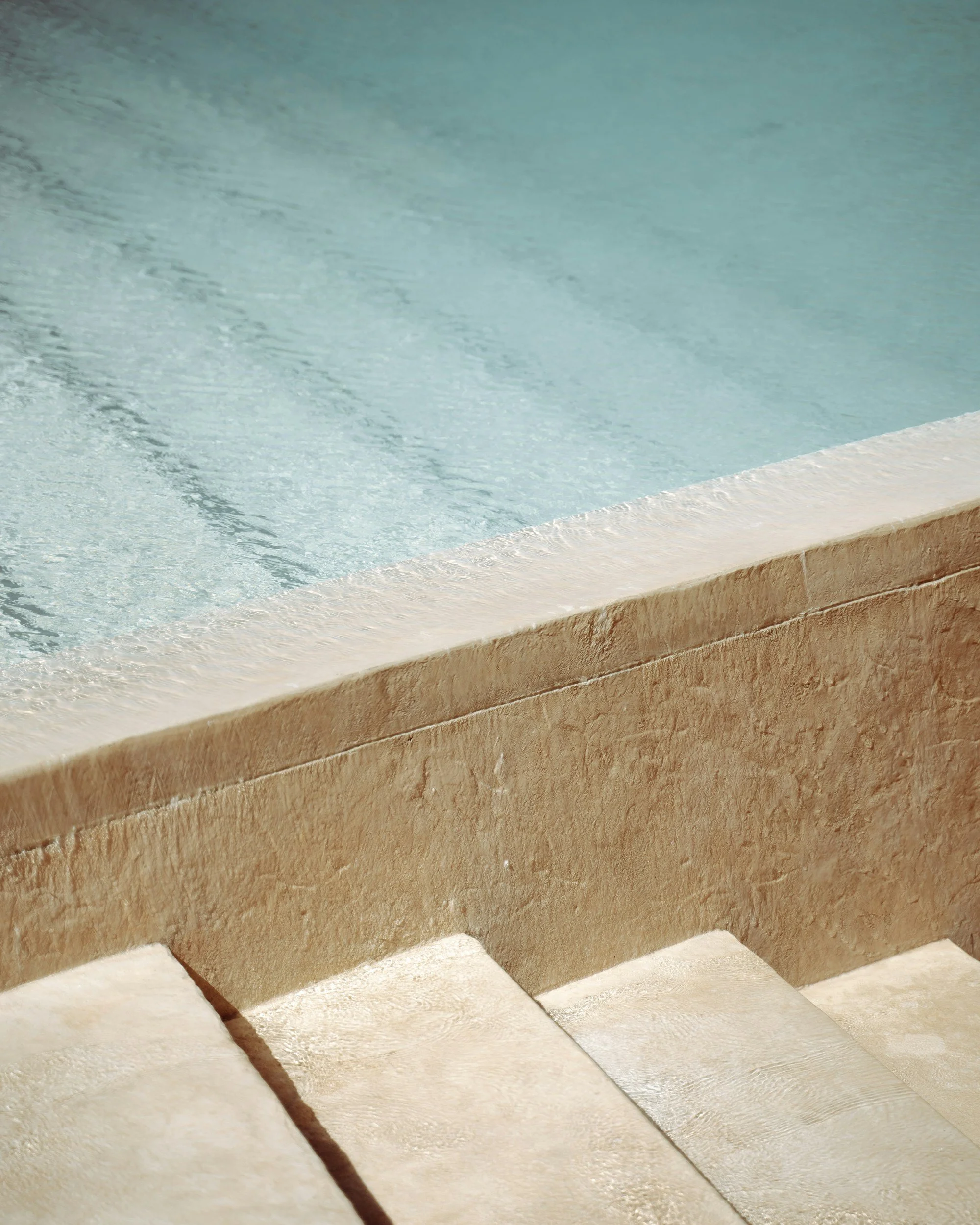 Close-up of an outdoor swimming pool with steps leading into the water, featuring a beige stone edge and a tiled pool bottom.