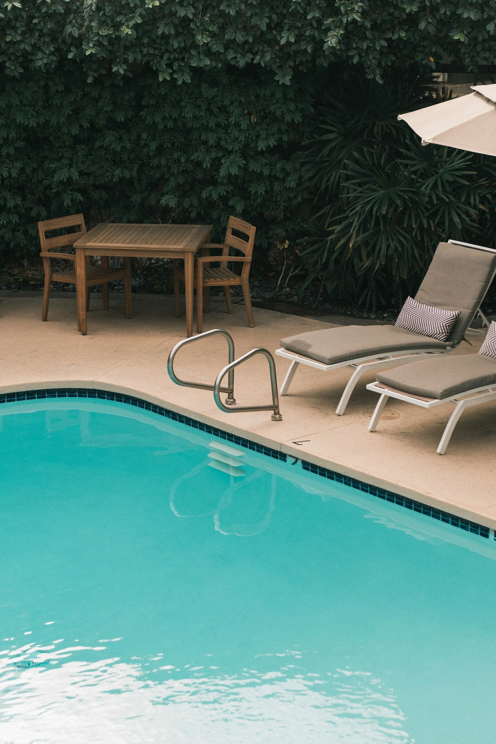 Swimming pool area with two lounge chairs, a small outdoor table and chairs, and surrounded by lush green foliage.