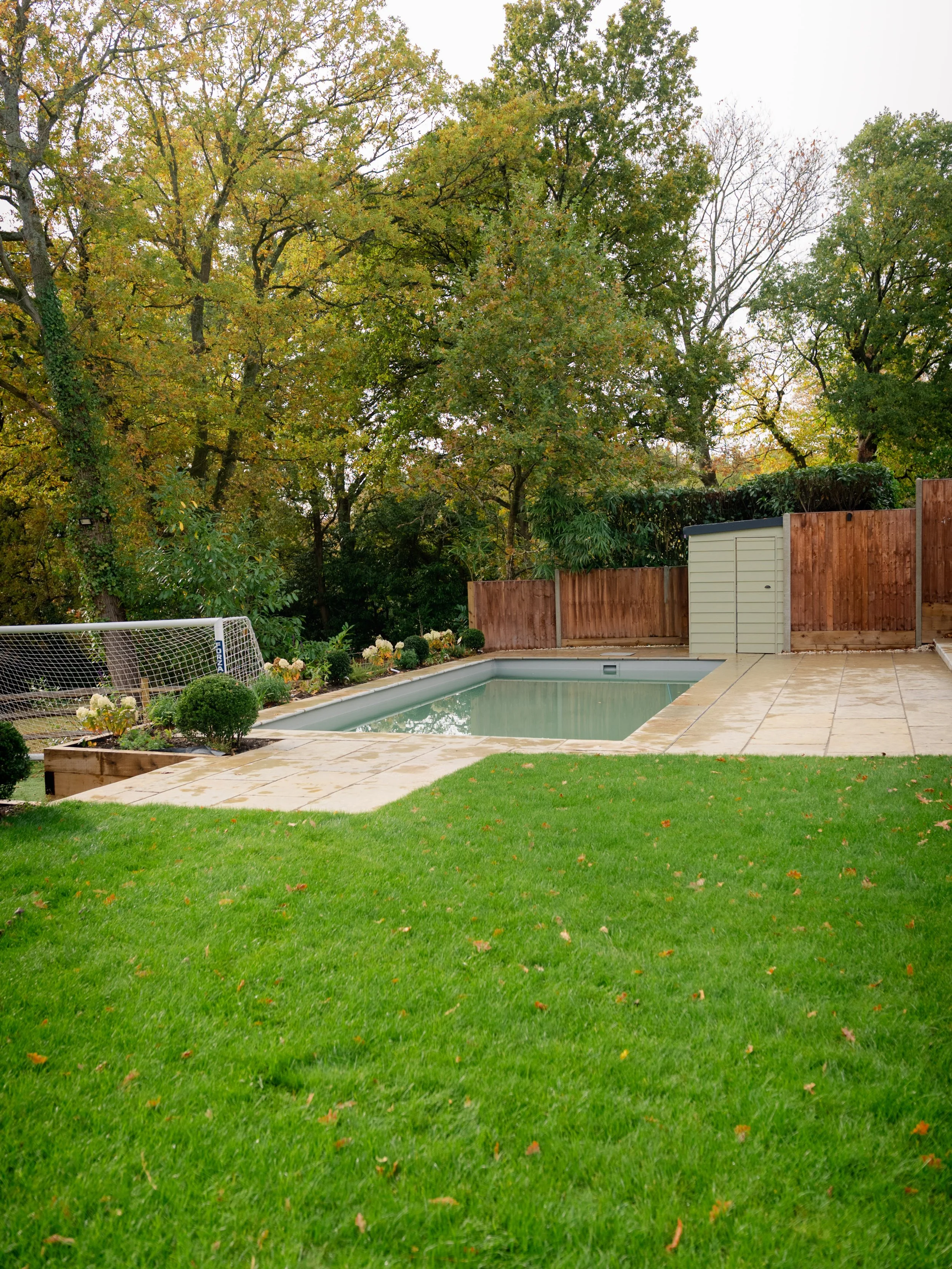 Backyard with a small swimming pool, surrounded by a paved patio, a grassy lawn, and a wooden fence with trees in the background.