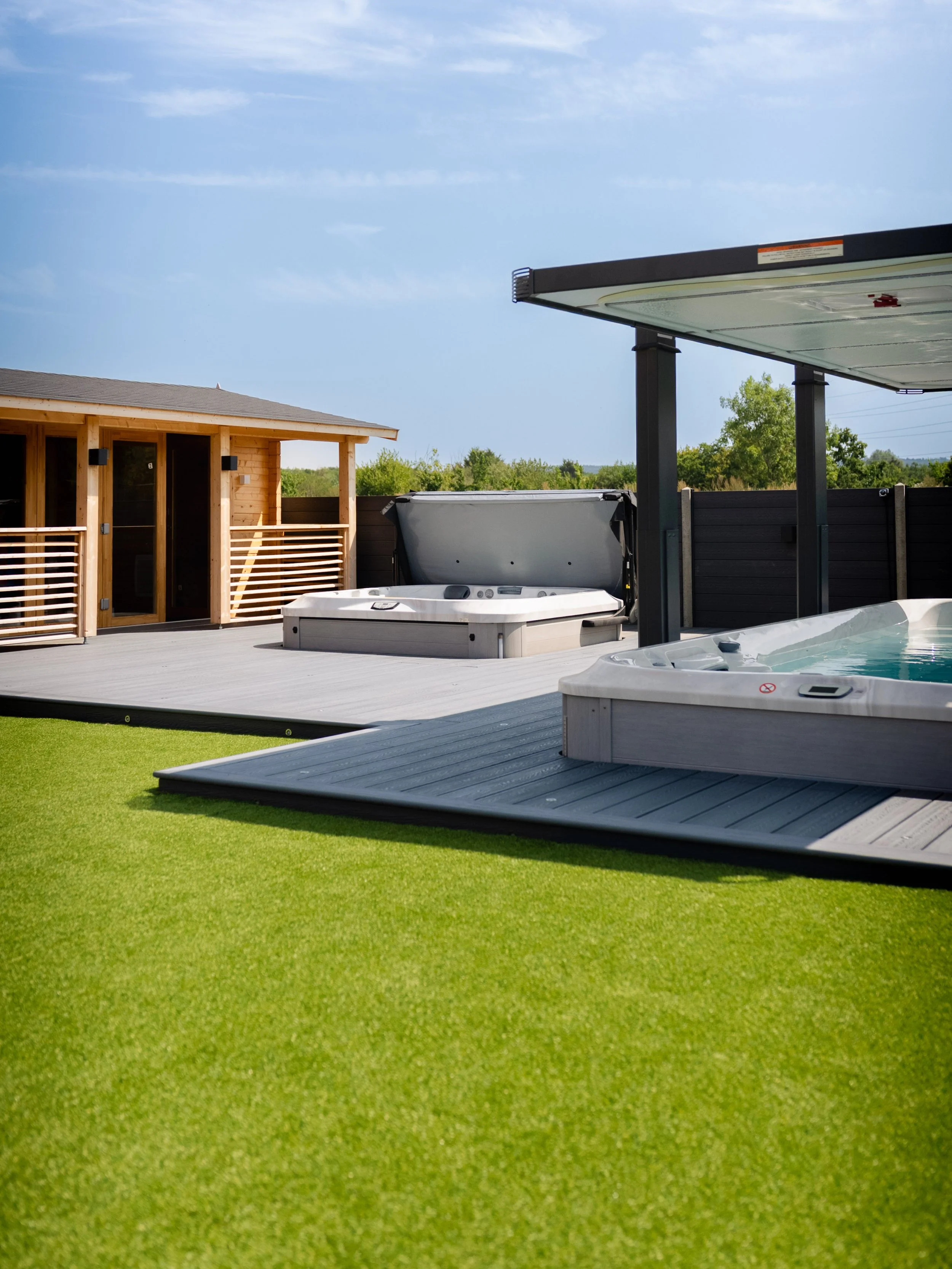 Outdoor backyard with hot tubs on a wooden deck, a wooden shed, and a covered area with a hot tub, surrounded by green grass and a privacy fence.
