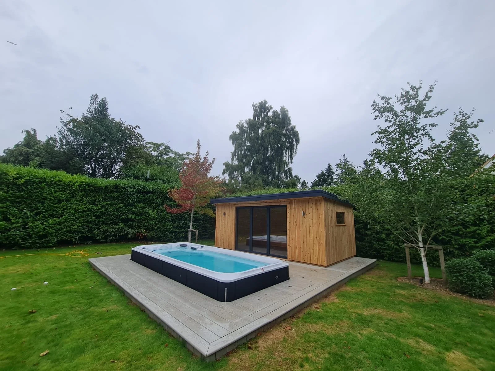 A backyard with a hot tub and a small wooden shed, surrounded by green trees and a hedge, on a cloudy day.