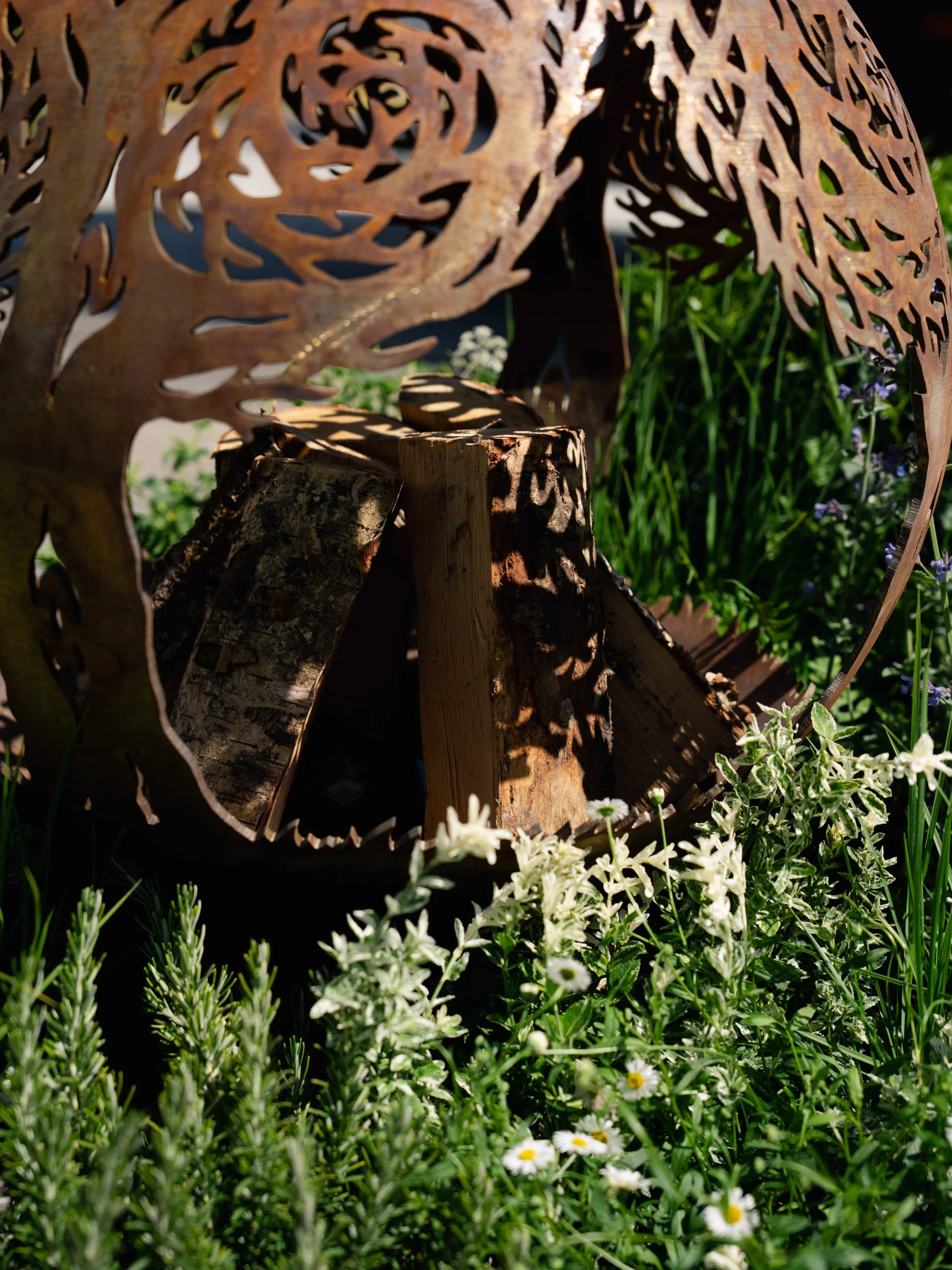 Rusty metal sculpture of a tree with intricate cutouts, partially surrounded by green plants and small white flowers.