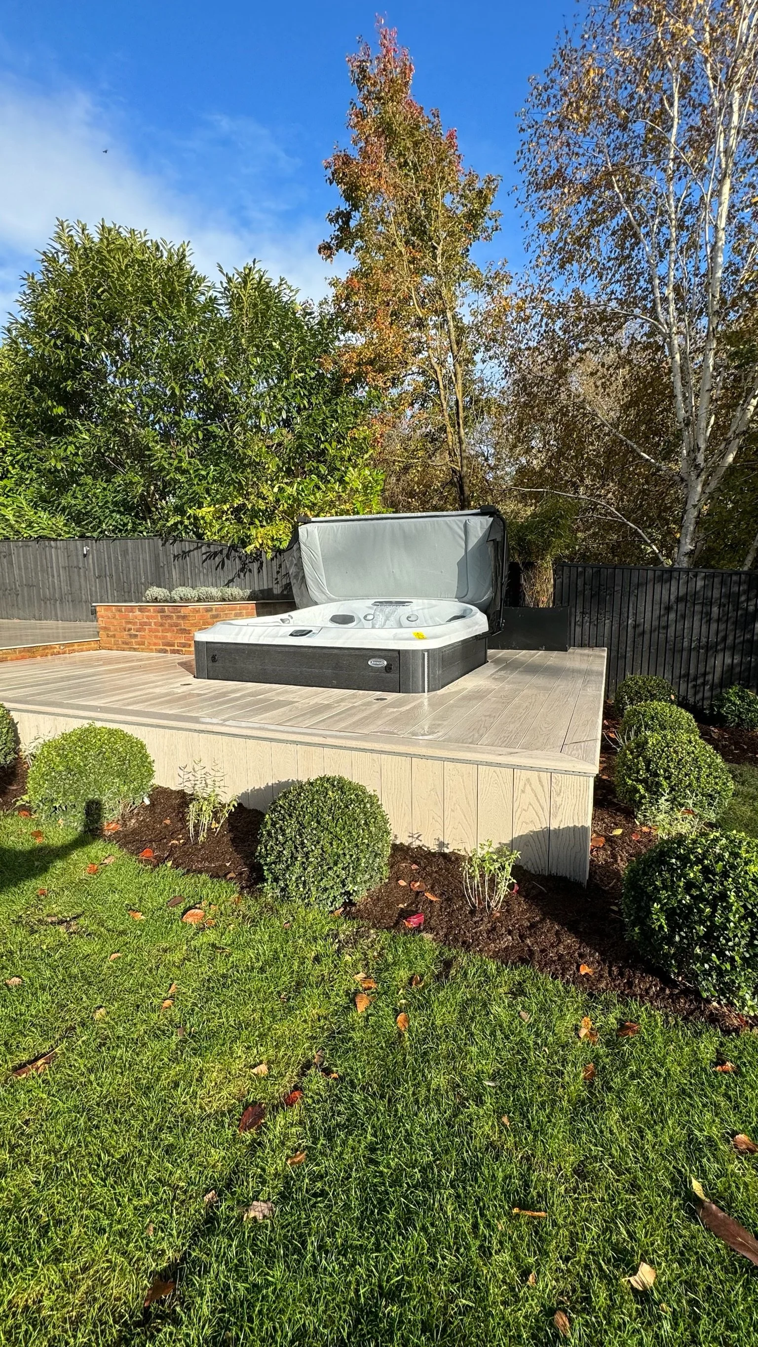 An outdoor hot tub on a wooden platform surrounded by landscaped bushes and grass, with trees and a fence in the background under a blue sky with some clouds.