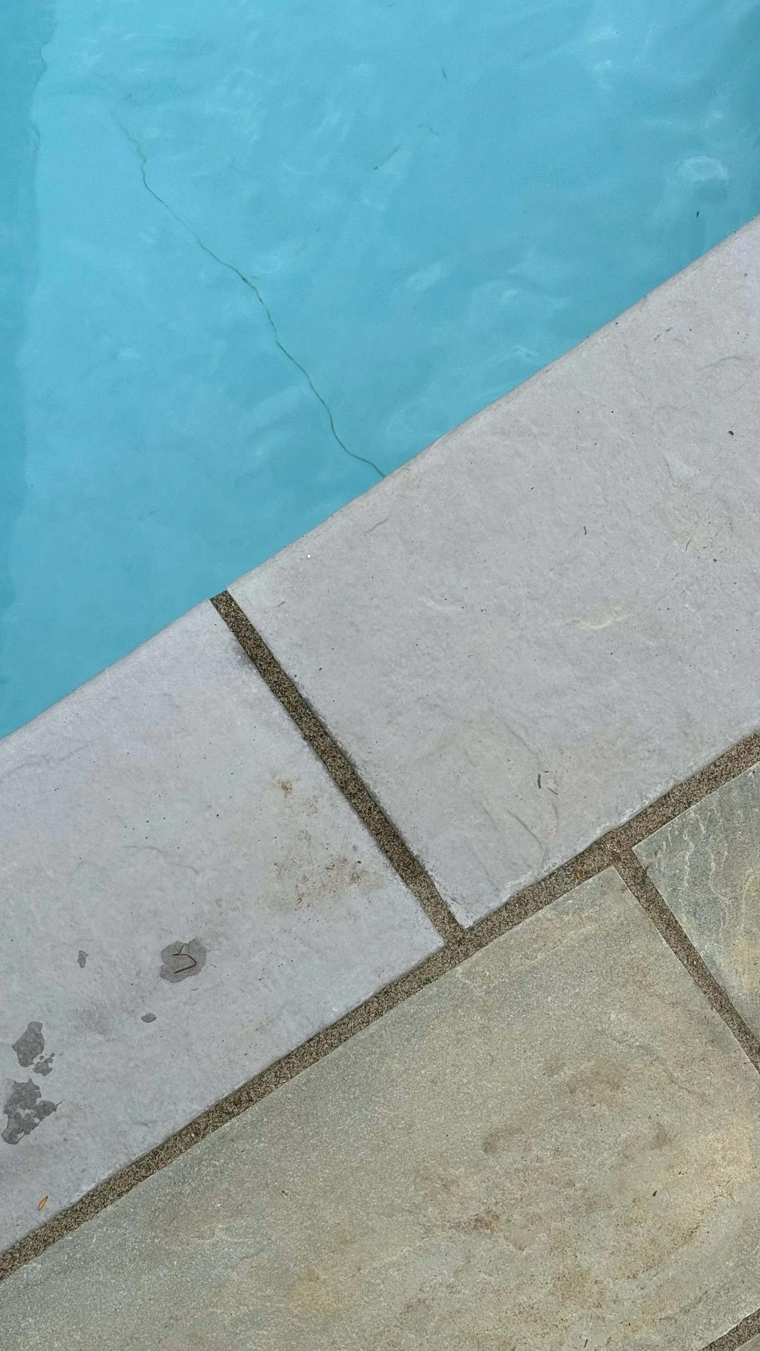 Close-up of a swimming pool corner with cracked light blue pool wall, beige tile and grout, and beige pool deck with dirt spots.