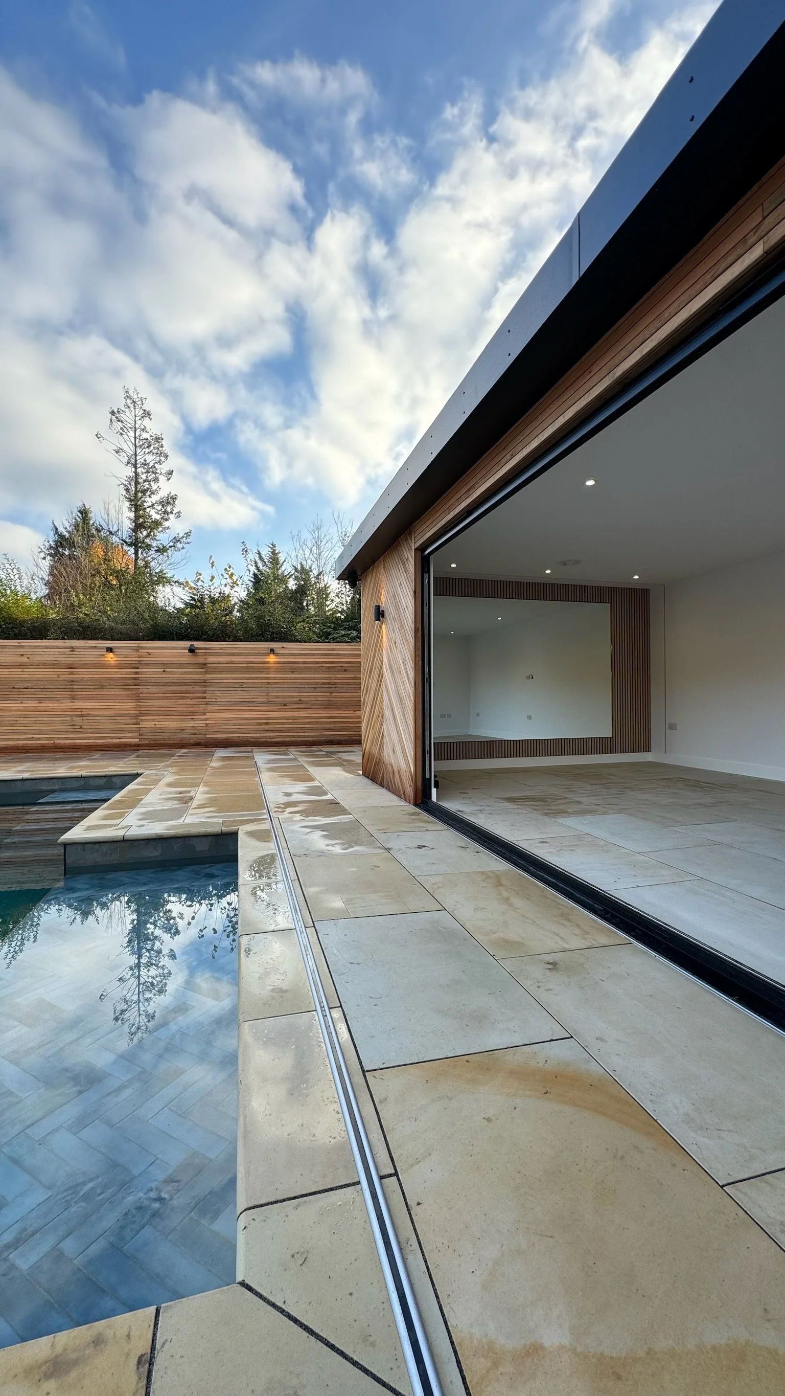 Modern house with large sliding glass doors open to a backyard with a swimming pool, stone patio, and wooden privacy fence. The sky is partly cloudy.