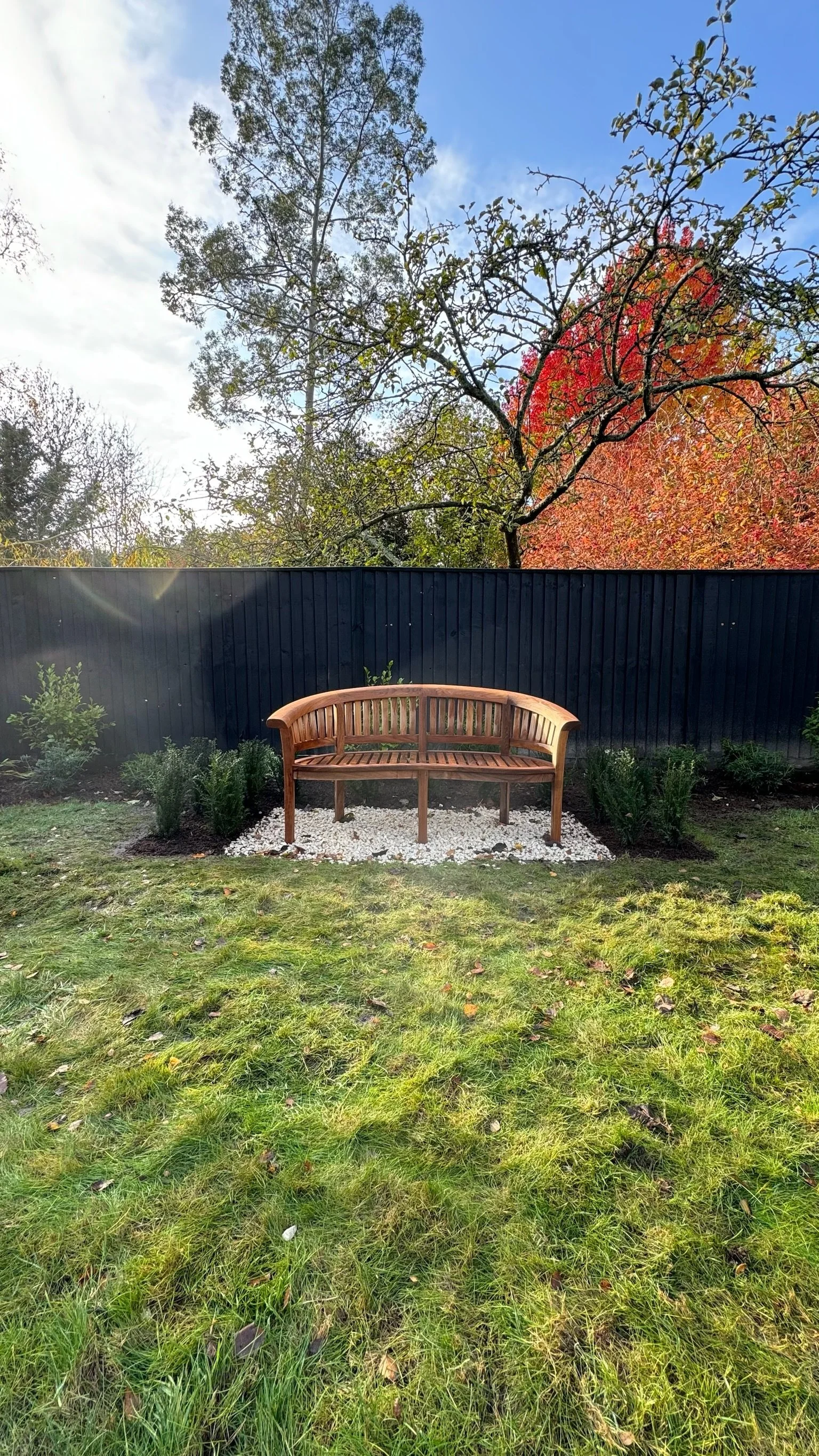 A garden with a wooden bench on a gravel patch, surrounded by green grass and small bushes. Esme are a widely trusted garden design and landscaping company serving surrey, hampshire, berkshire, ascot, london and surrounding areas.