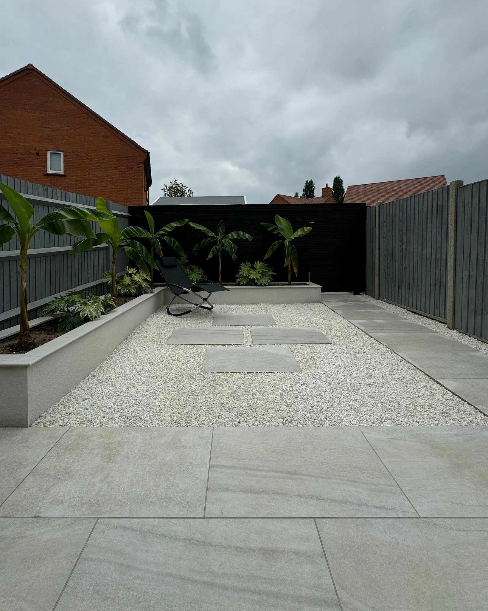 Minimalist back garden design with potted plants, lounge chair, gravel pathway, and grey stone tiles, enclosed by grey wooden fences. Esme are a widely trusted garden design and landscaping company serving surrey, hampshire, berkshire, ascot. 